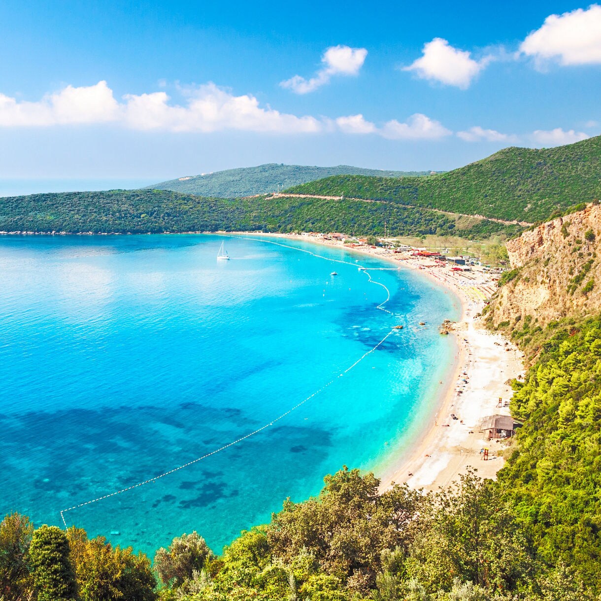 A crescent-shaped bay with turquoise water surrounded by green forested hills, with a sandy beach and sailboat visible in the distance.