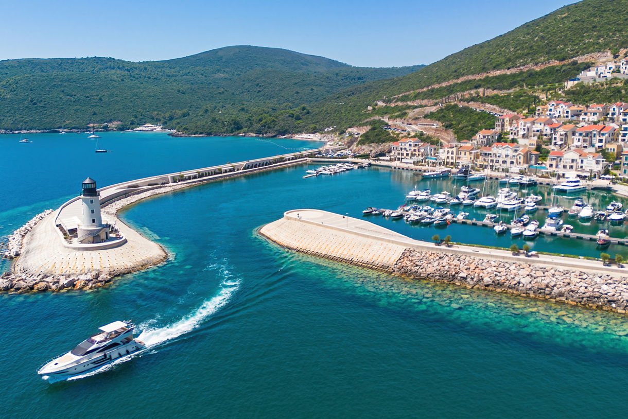 An aerial view shows a white luxury yacht cruising toward a sheltered marina surrounded by turquoise blue water. A small lighthouse stands on the end of a curved breakwater guiding boats into the harbor. Numerous sailboats and motorboats are docked along the piers beside hillside houses and apartments. Green hills rise in the background under a clear, sunny sky, creating a relaxed coastal vacation atmosphere.