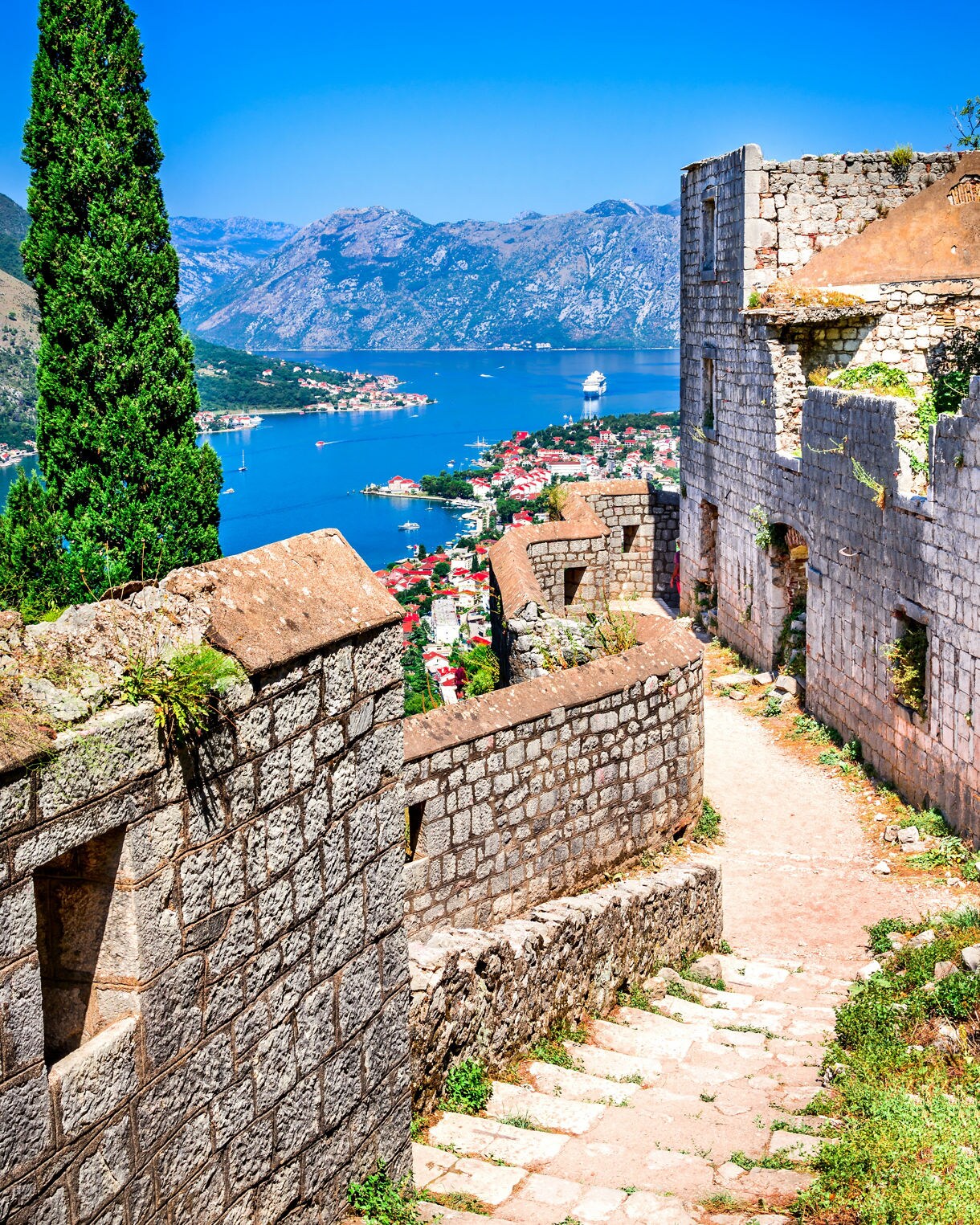 Stone fortress walls and pathway overlooking the Bay of Kotor, with red-roofed houses, blue sea and surrounding mountains.