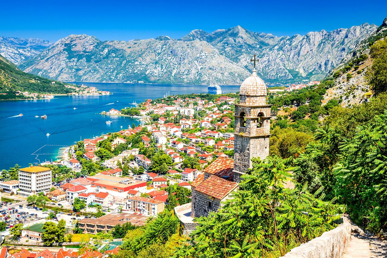 Scenic view over Kotor, Montenegro, with a stone church tower in the foreground, red-roofed buildings along the bay and dramatic mountains rising in the background.