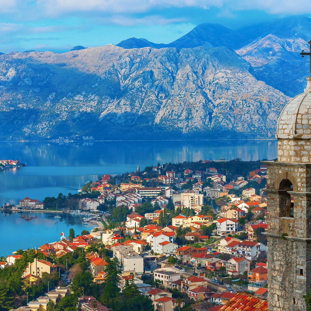 A stone church bell tower with a domed roof and cross overlooking the red-roofed town of Kotor beside a calm blue bay and rugged mountains.