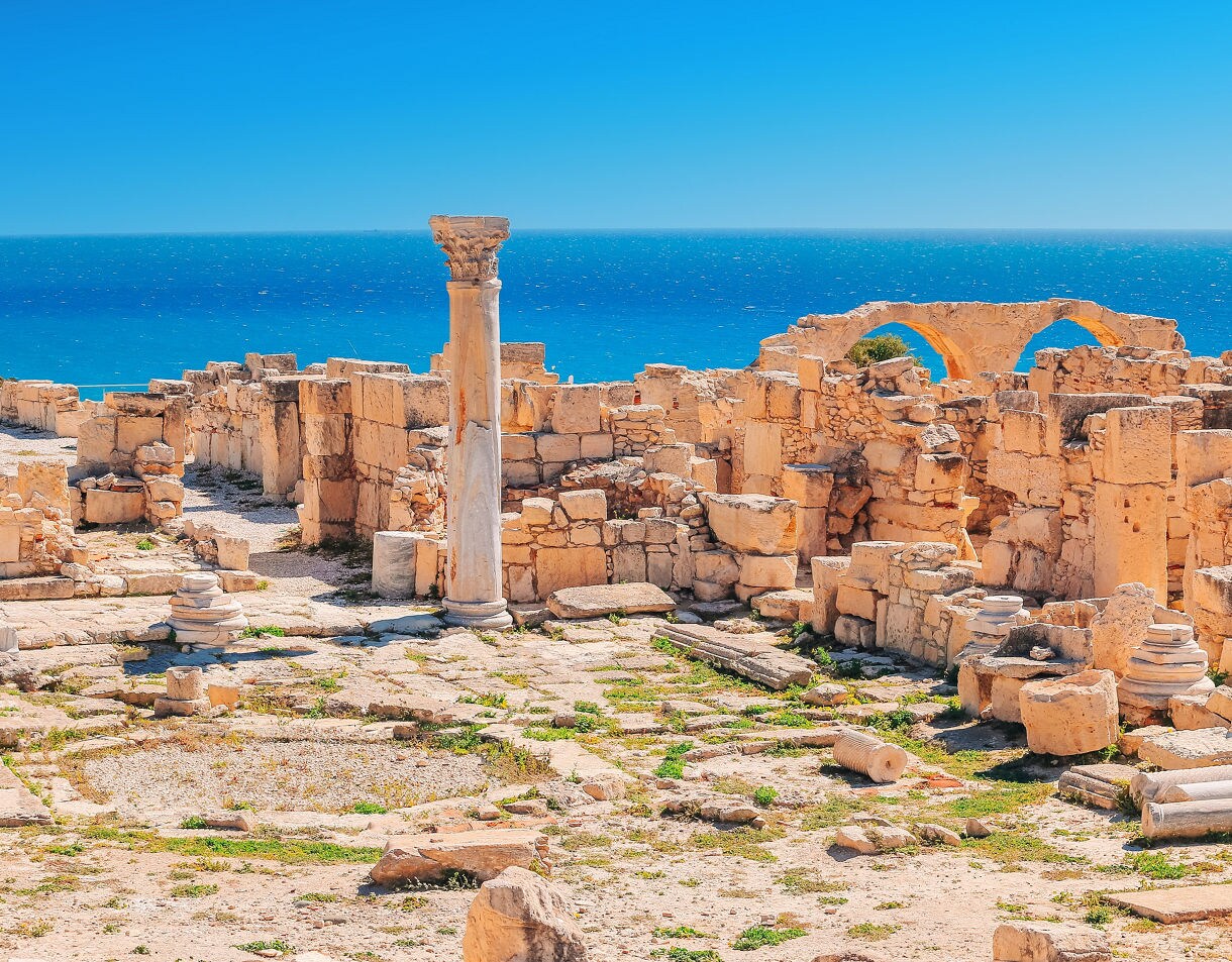 Ruins of the Kourion archaeological site overlooking bright blue Mediterranean waters with broken columns and stone walls scattered across the hillside.