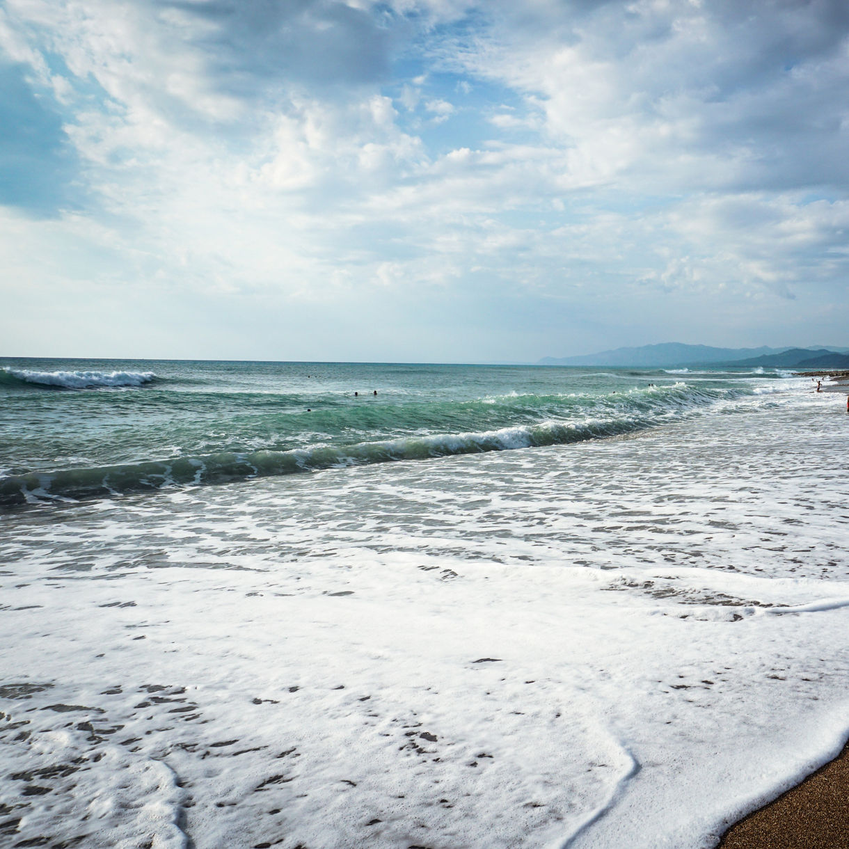 Wide view of Kourouta Beach in Greece with gentle waves washing over golden sand and people swimming and relaxing along the shore under a partly cloudy sky.