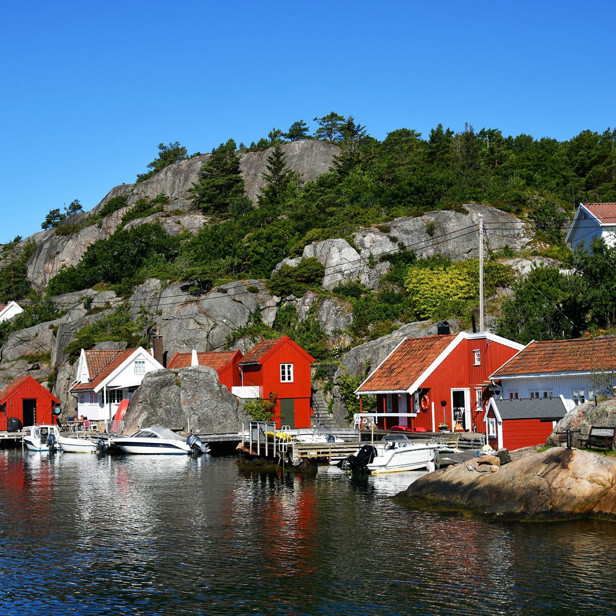 Small red and white wooden houses along a rocky shoreline with boats tied to docks and pine-covered hills rising behind.