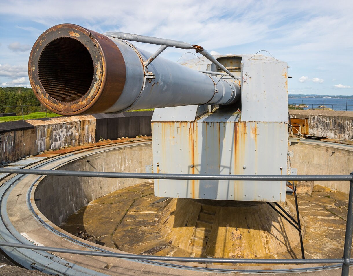 A large rusting coastal artillery cannon mounted in a concrete fortification with ocean and trees visible in the background.