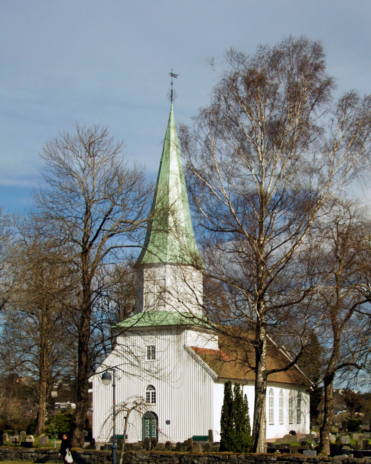A white wooden church with a tall green steeple surrounded by leafless trees and a small graveyard under a clear sky.