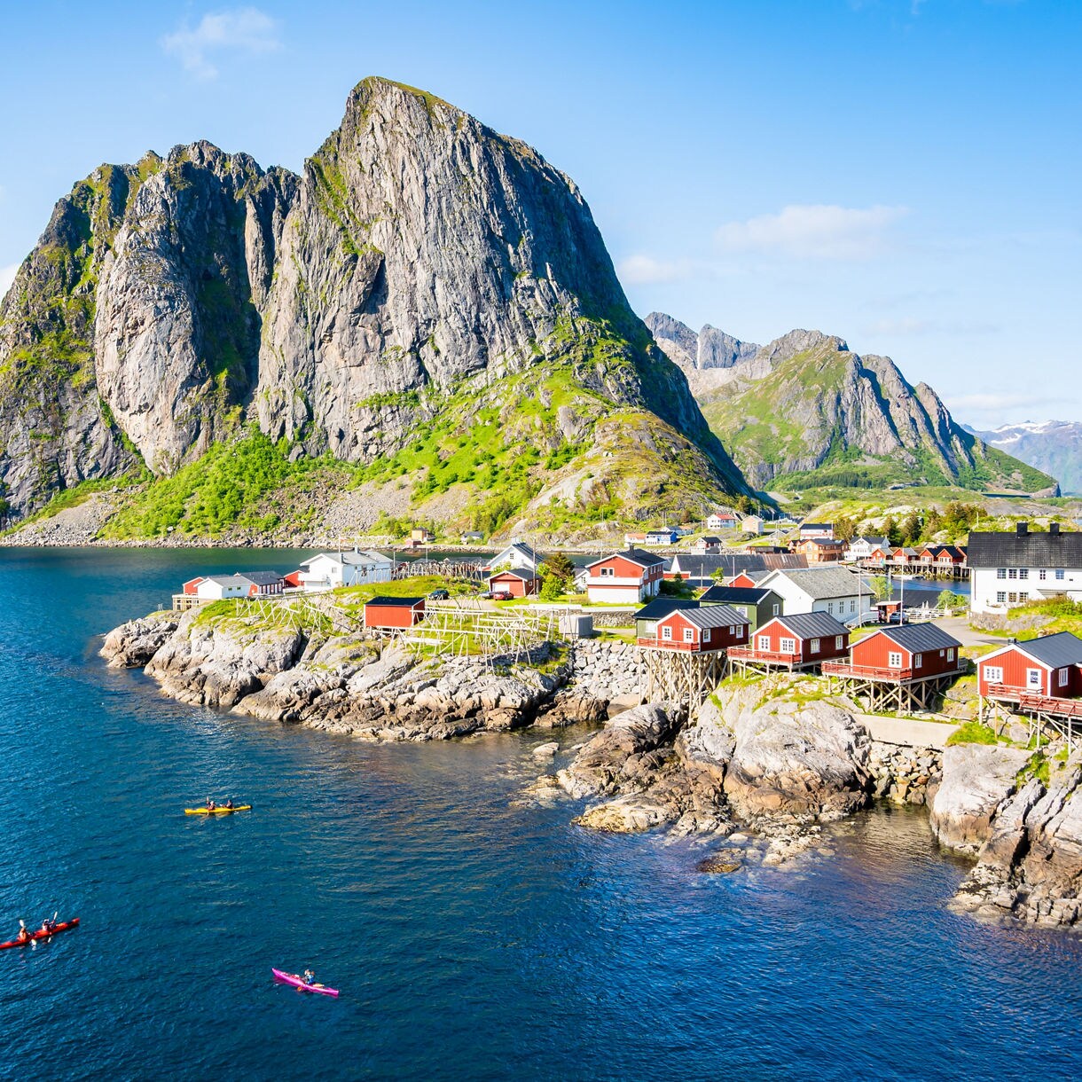 Kayakers in calm blue water near red coastal cabins built on rocky cliffs, with steep green mountains rising behind under a clear sky.