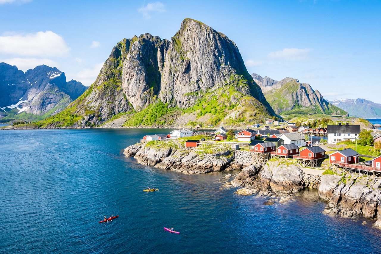 A coastal Norwegian village with red and white wooden houses lining a sheltered inlet, small boats tied to docks and rocky hills in the background.