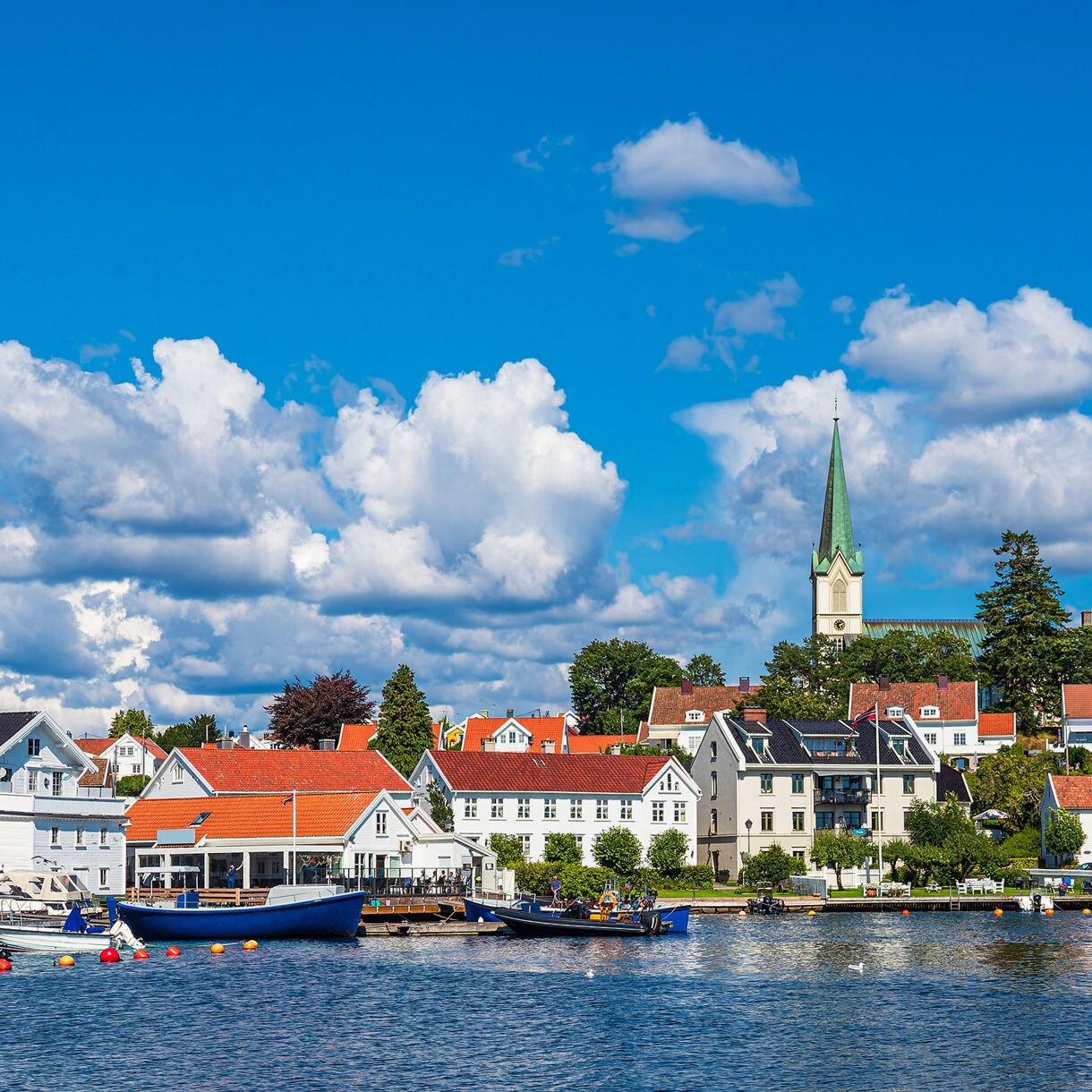 A coastal Norwegian town with white and red-roofed houses, a tall green church steeple, and small boats floating on calm blue water under a clear sky with fluffy clouds.