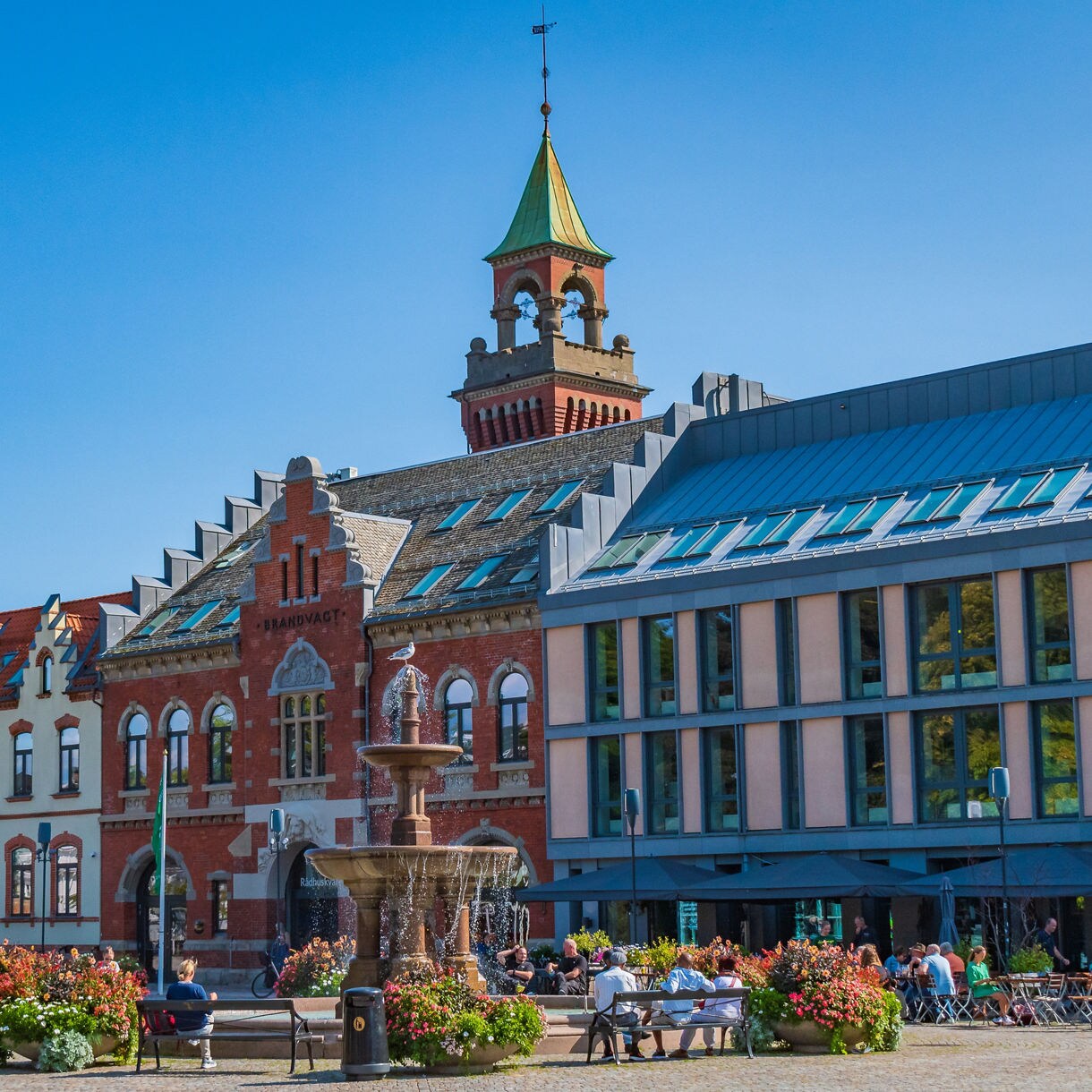 A historic European town square with brick and pastel buildings, a central fountain, flower planters, and people sitting at outdoor tables under a clear blue sky.