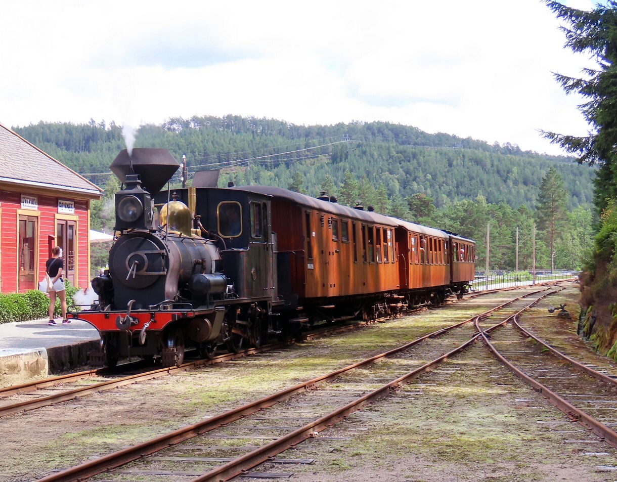 A vintage steam locomotive with wooden passenger cars stopped at a small rural station, surrounded by green forested hills.