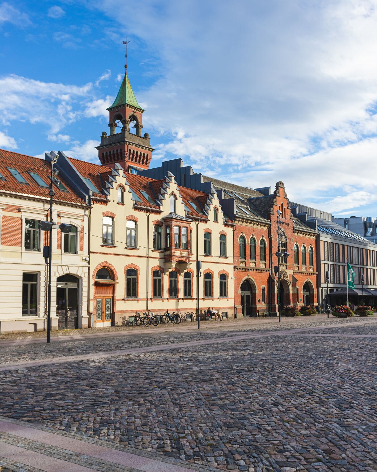 A cobblestone plaza bordered by decorative brick and cream-colored buildings with a tall green-roofed tower rising above under a partly cloudy sky.