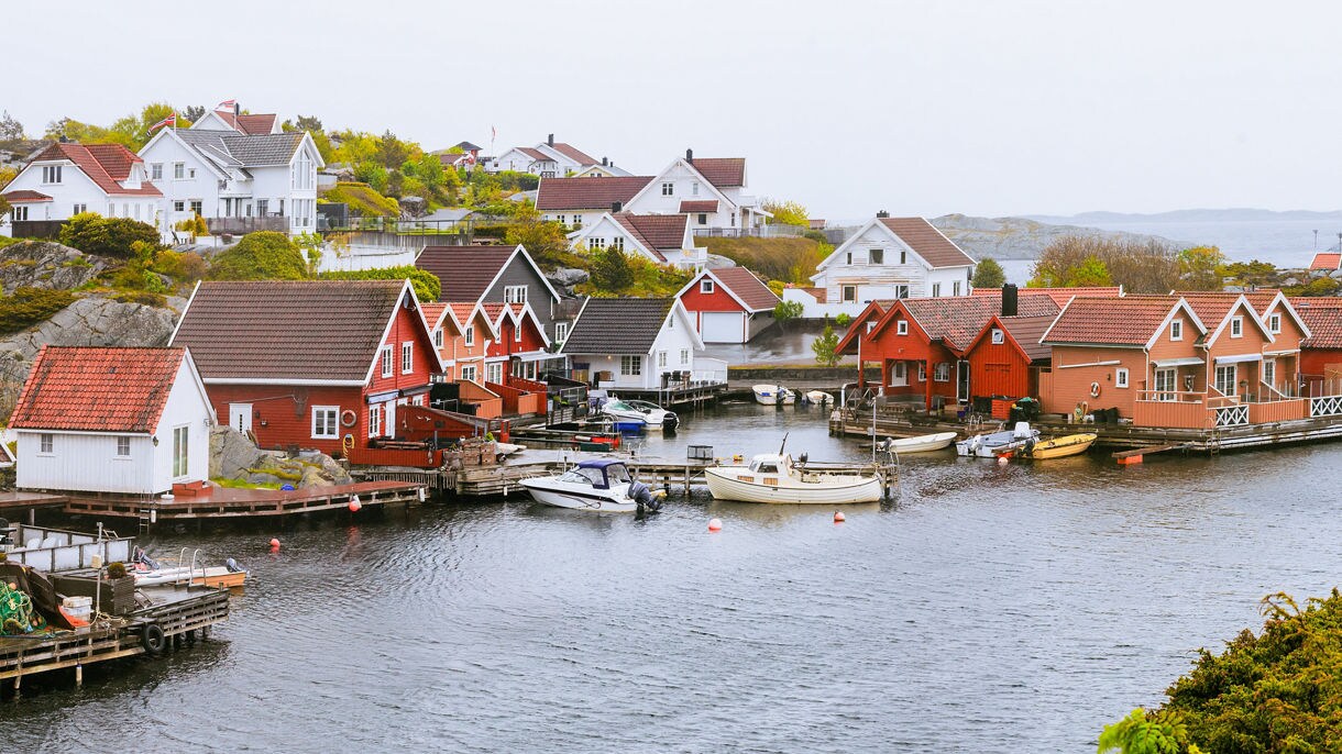 A coastal Norwegian village with red and white wooden houses lining a sheltered inlet, small boats tied to docks and rocky hills in the background.