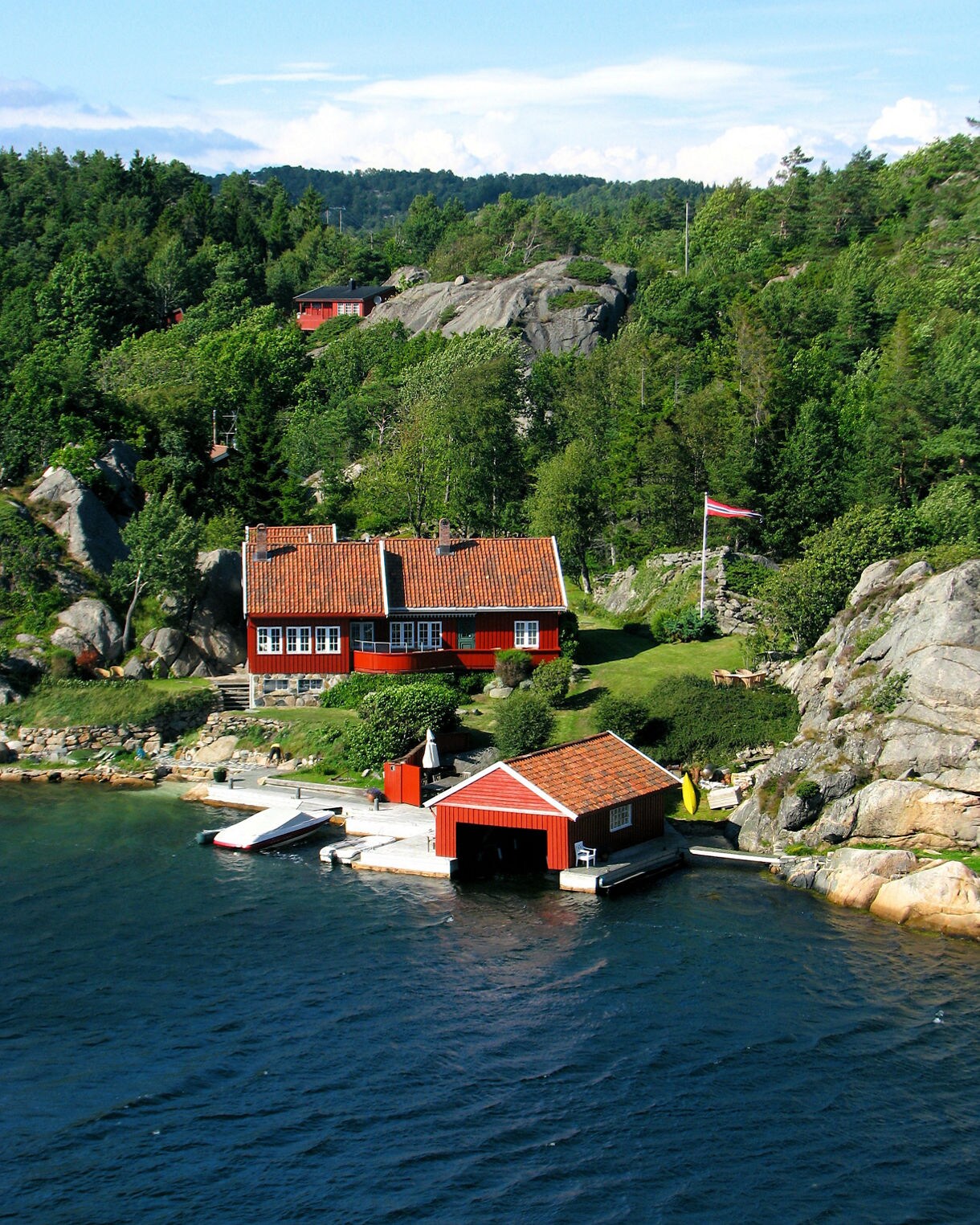 A red waterfront cabin and boathouse nestled among rocky hills and dense green trees with small docks and boats along the shoreline.