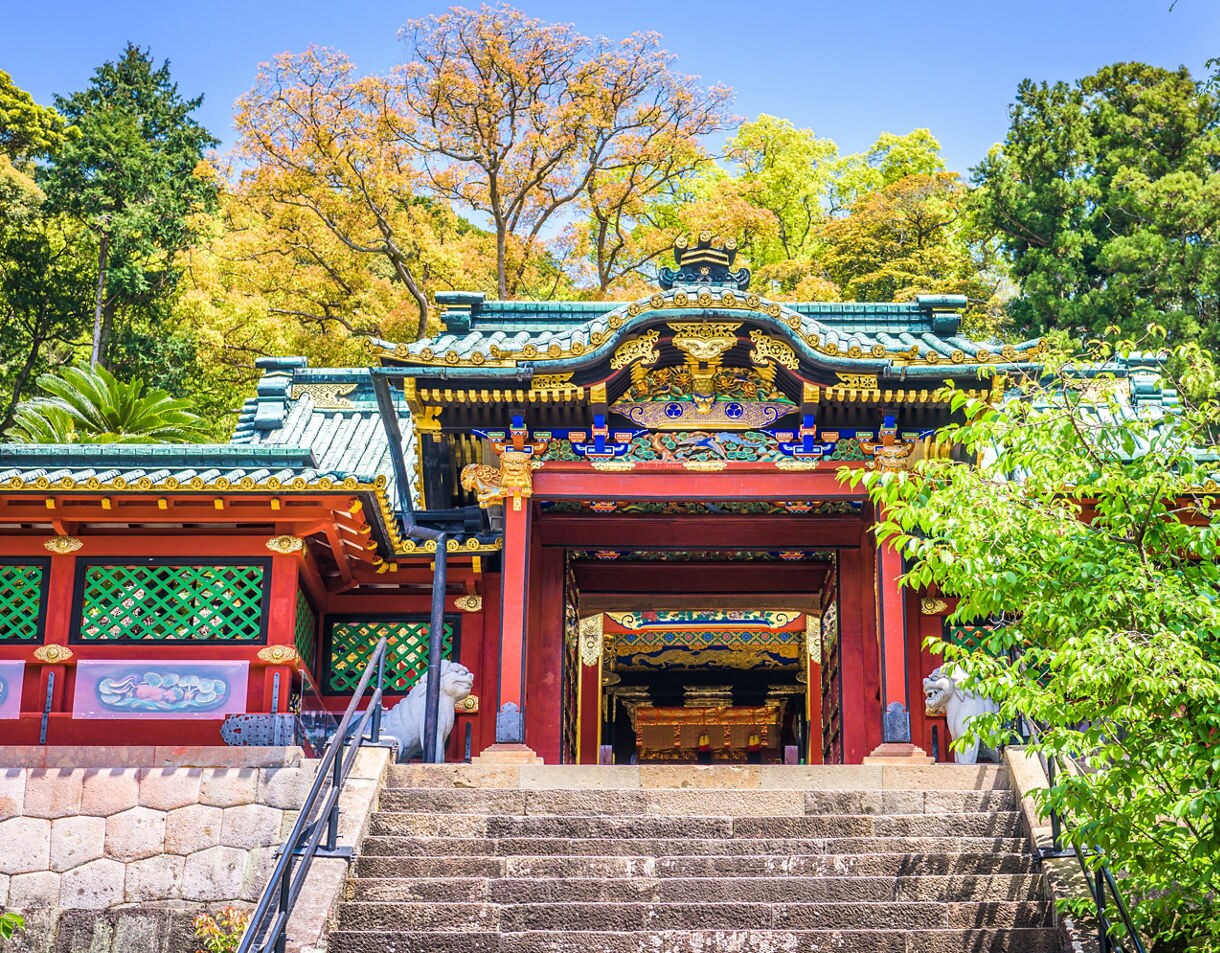 Brightly decorated entrance of a traditional Japanese shrine with red pillars, green lattice panels and intricate gold details, set atop stone steps and framed by lush trees under a clear sky.