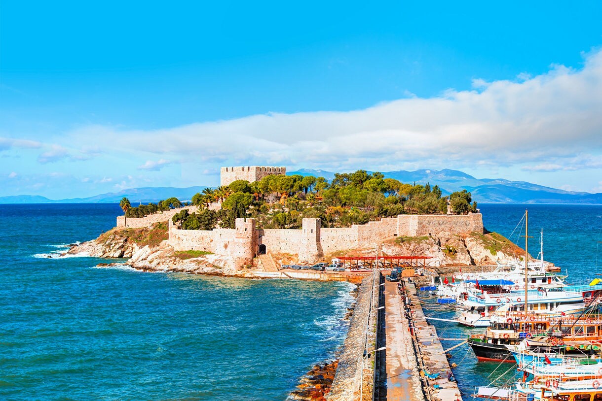 A stone causeway leading to a small island with a historic fortress surrounded by trees, with colorful boats docked along the shore and deep blue sea all around.