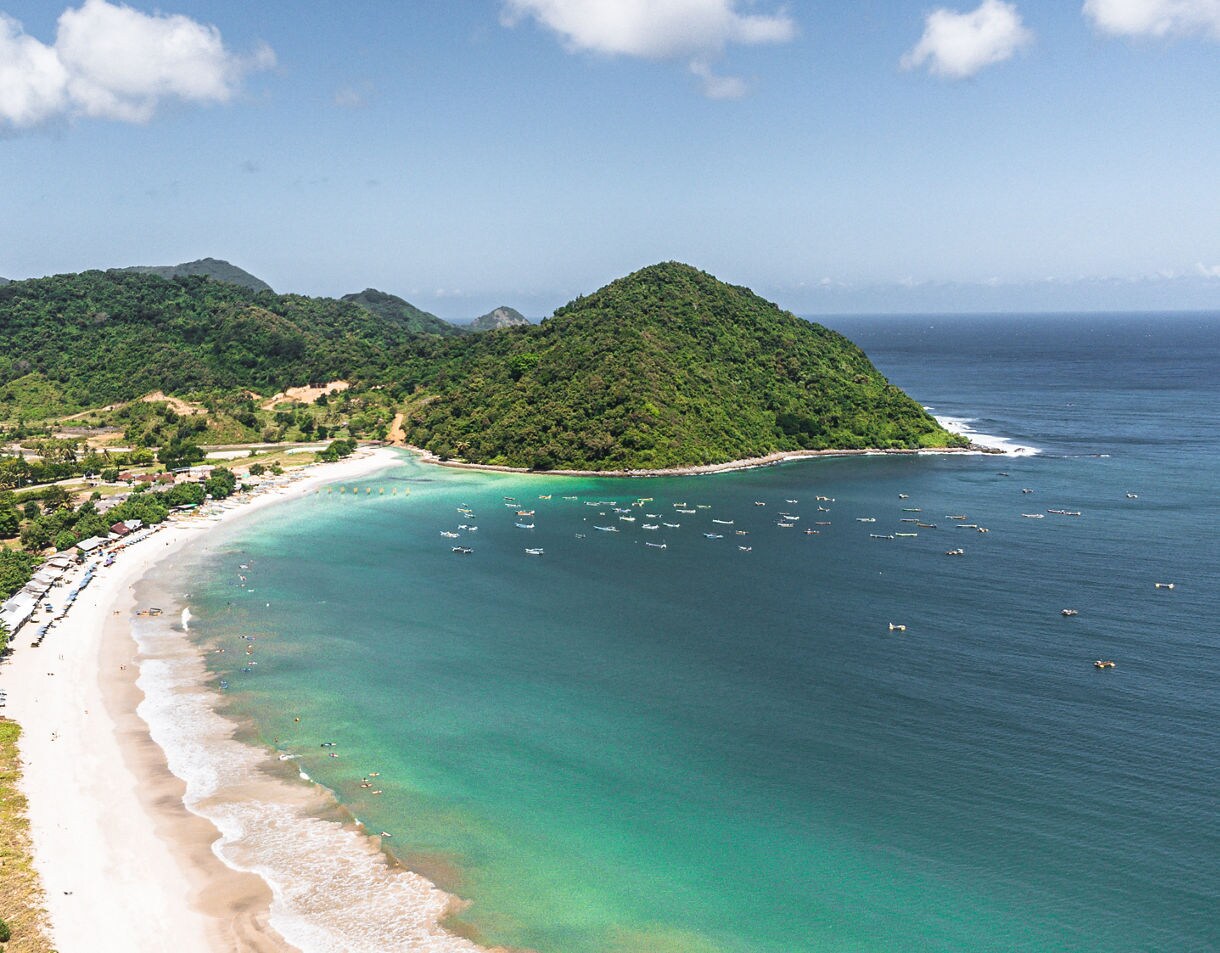 Aerial view of a sweeping tropical bay with clear turquoise shallows, white sand beach, clusters of small boats on deep blue water and lush green hills rising behind the shoreline.