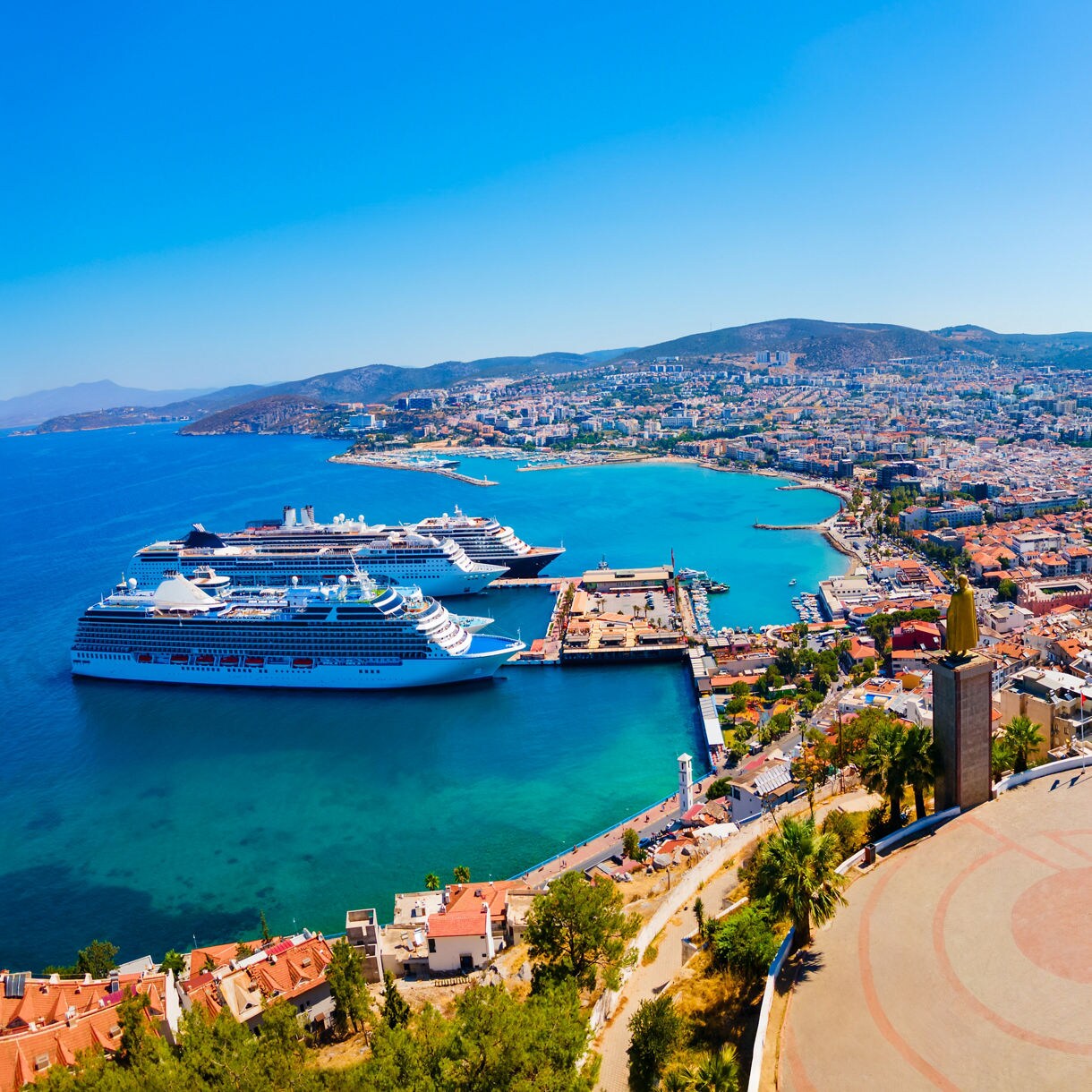 Panoramic view of Kuşadası, Turkey, showing a bright blue harbor with cruise ships docked, red-roofed buildings across the shoreline and surrounding hills.
