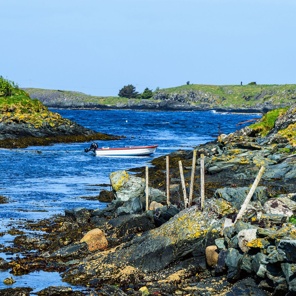 A small white boat floats on vivid blue water surrounded by rocky, seaweed-covered shores and grassy islets along Norway’s coastal trail.