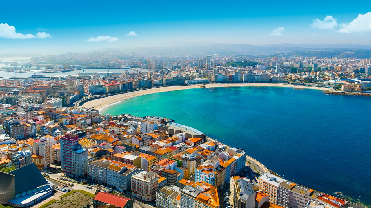 Aerial view of A Coruña, Spain, showing colorful city buildings, a wide sandy beach and a curved blue bay under a clear sky.