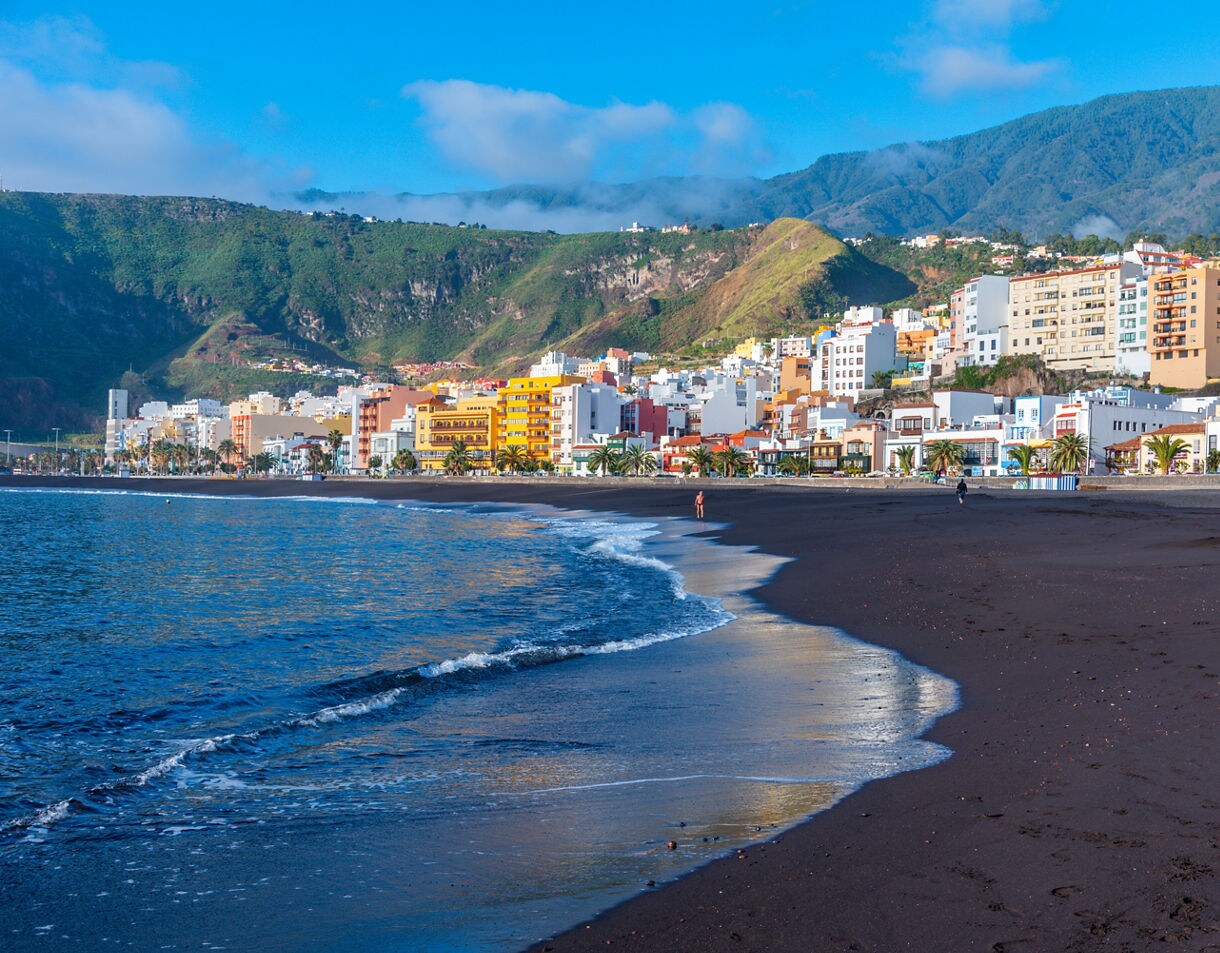 Black sand beach along the coast of Santa Cruz de La Palma with gentle waves rolling onto shore and rows of colorful buildings backed by lush green mountains.