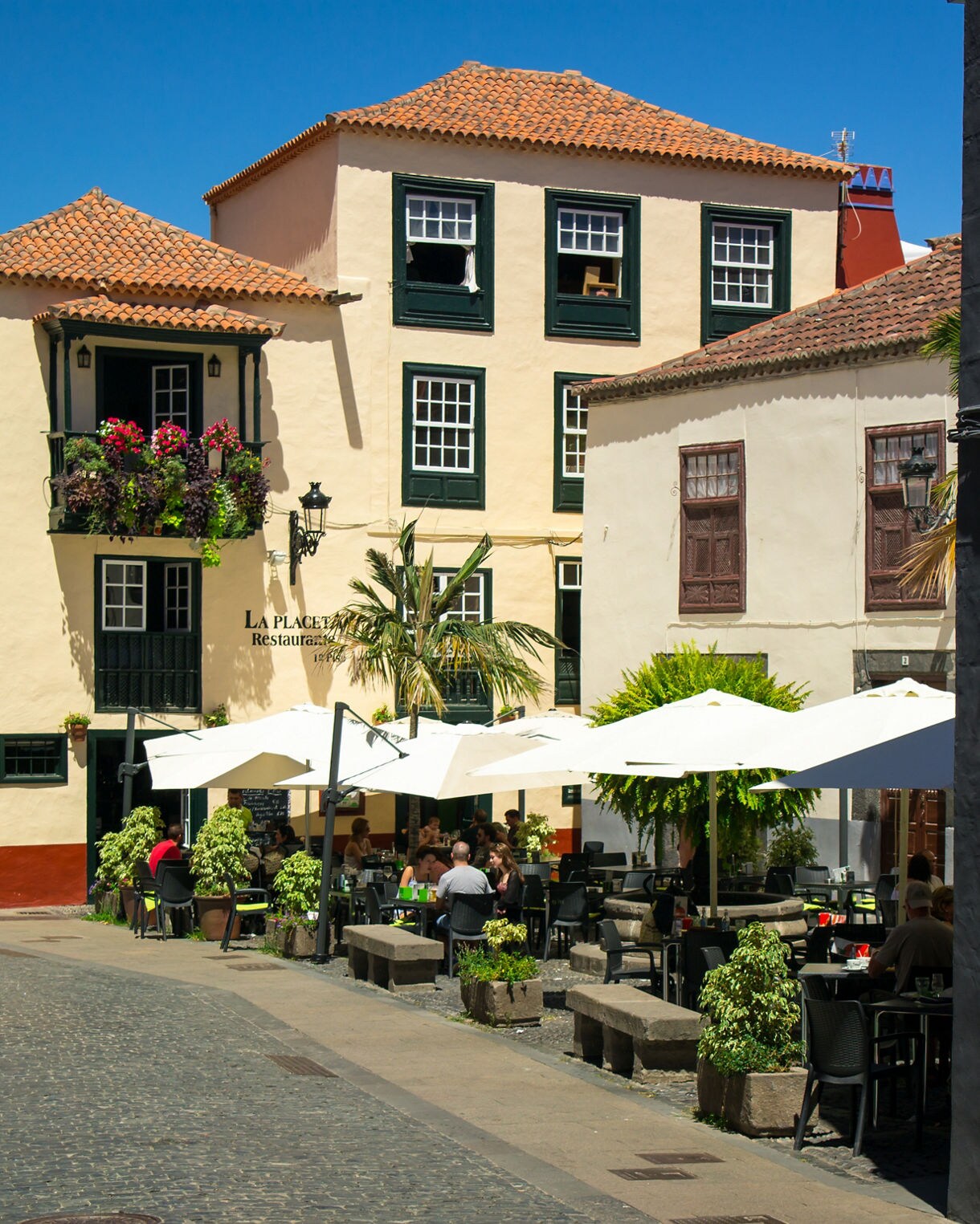 Cobblestone street in Santa Cruz de La Palma lined with colorful colonial buildings where people sit beneath white umbrellas at an outdoor café terrace.