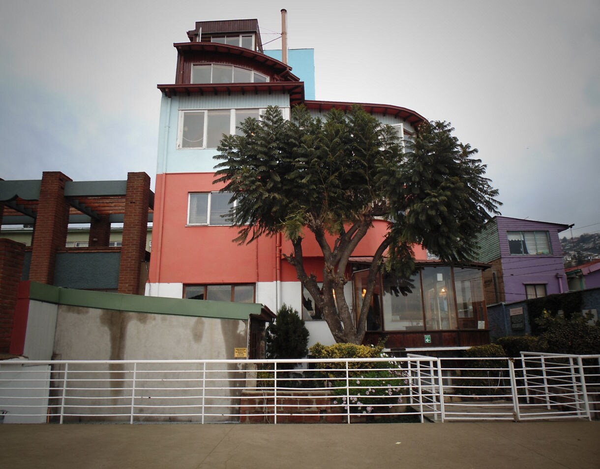 Multicolored house known as La Sebastiana in Valparaíso with tall windows, a large tree in front and surrounding hillside homes.