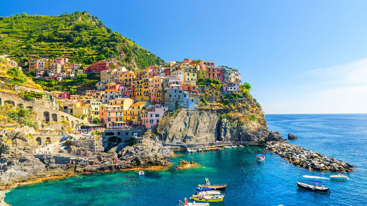 A colorful hillside village perched on rocky cliffs above clear turquoise sea, with small boats floating in the harbor and terraced green slopes rising behind.