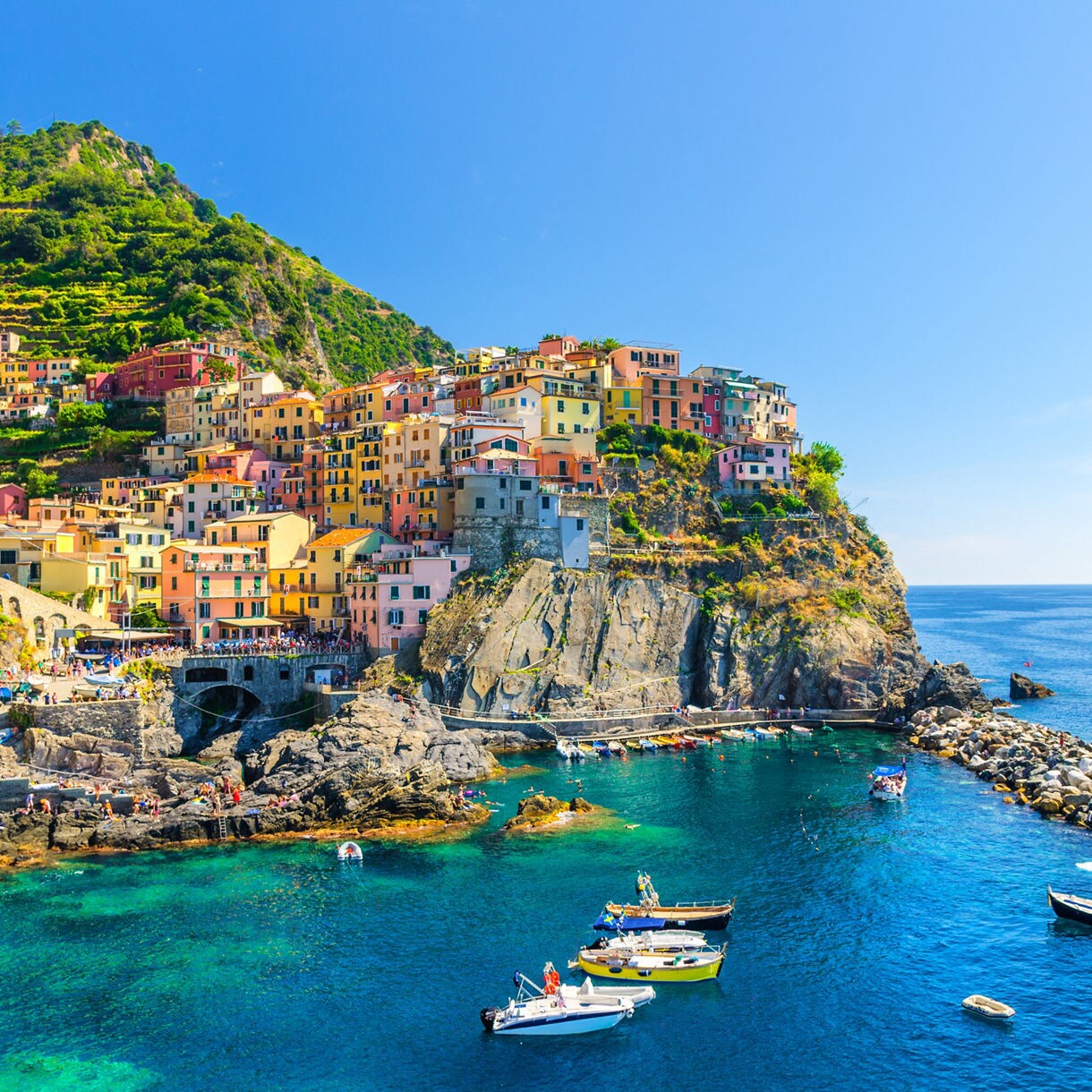 A colorful hillside village perched on rocky cliffs above clear turquoise sea, with small boats floating in the harbor and terraced green slopes rising behind.