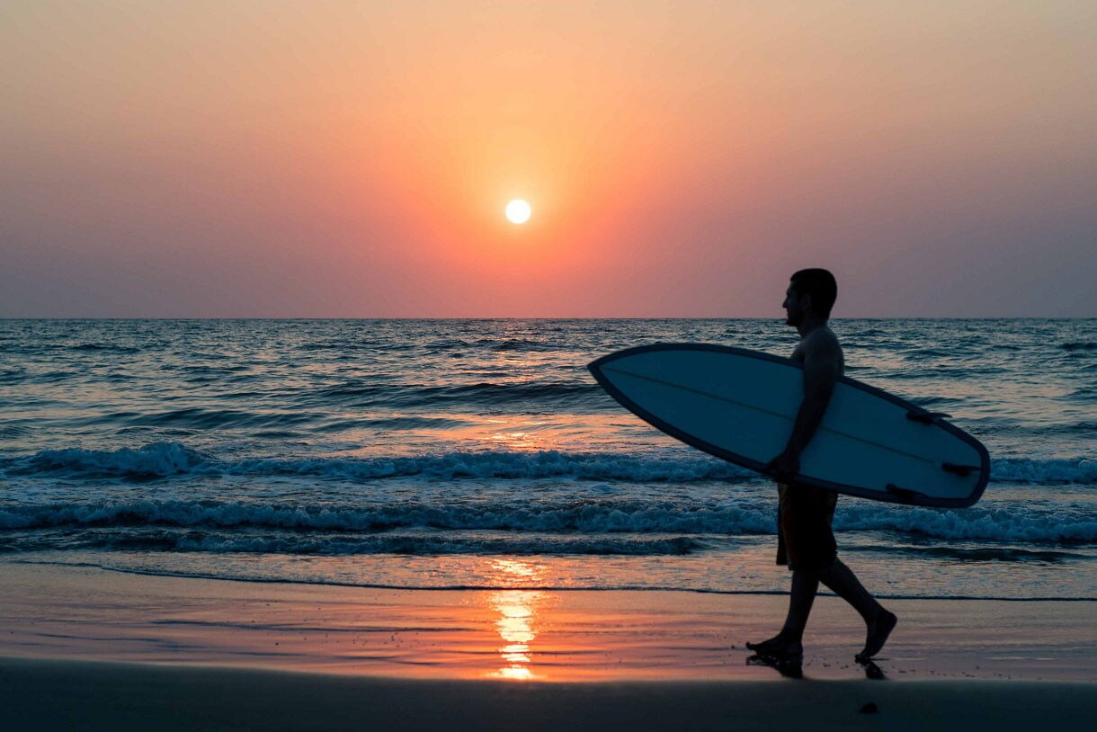 A surfer walks along the shoreline at La Jolla Shores, carrying a surfboard under one arm as the sun sets over the Pacific Ocean.