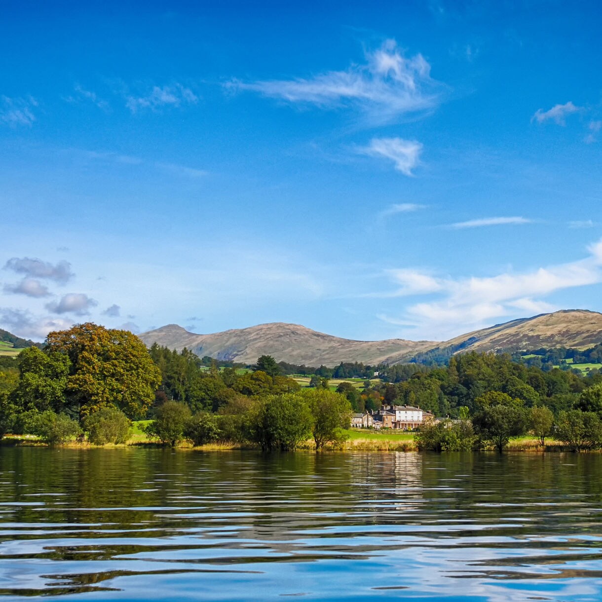 Scenic view of Lake Windermere with calm reflective water, rolling green hills, scattered trees and a distant house under a blue sky with wispy clouds.