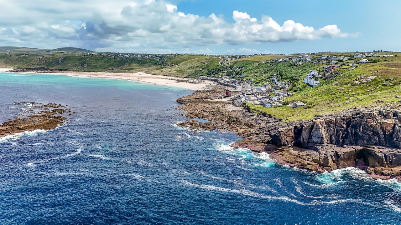 Aerial view of Land’s End in Cornwall with dramatic cliffs, a sandy beach, turquoise waters and scattered houses along the coast.