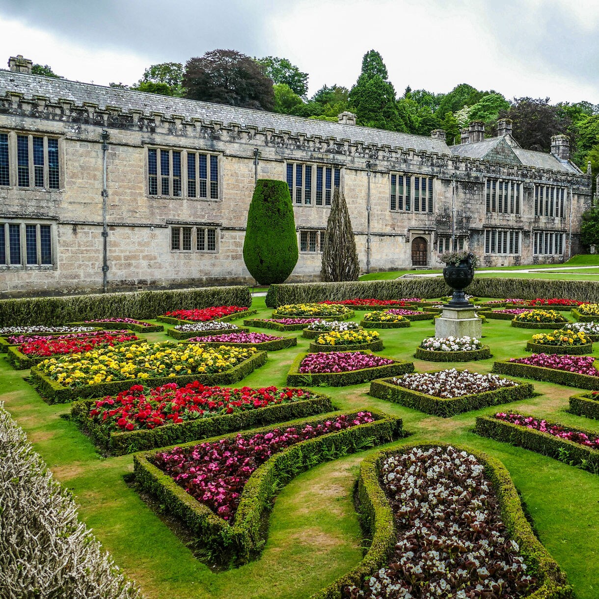 Formal gardens with colorful flower beds arranged in geometric patterns in front of Lanhydrock House, a stately stone manor with tall windows and an adjoining church tower.