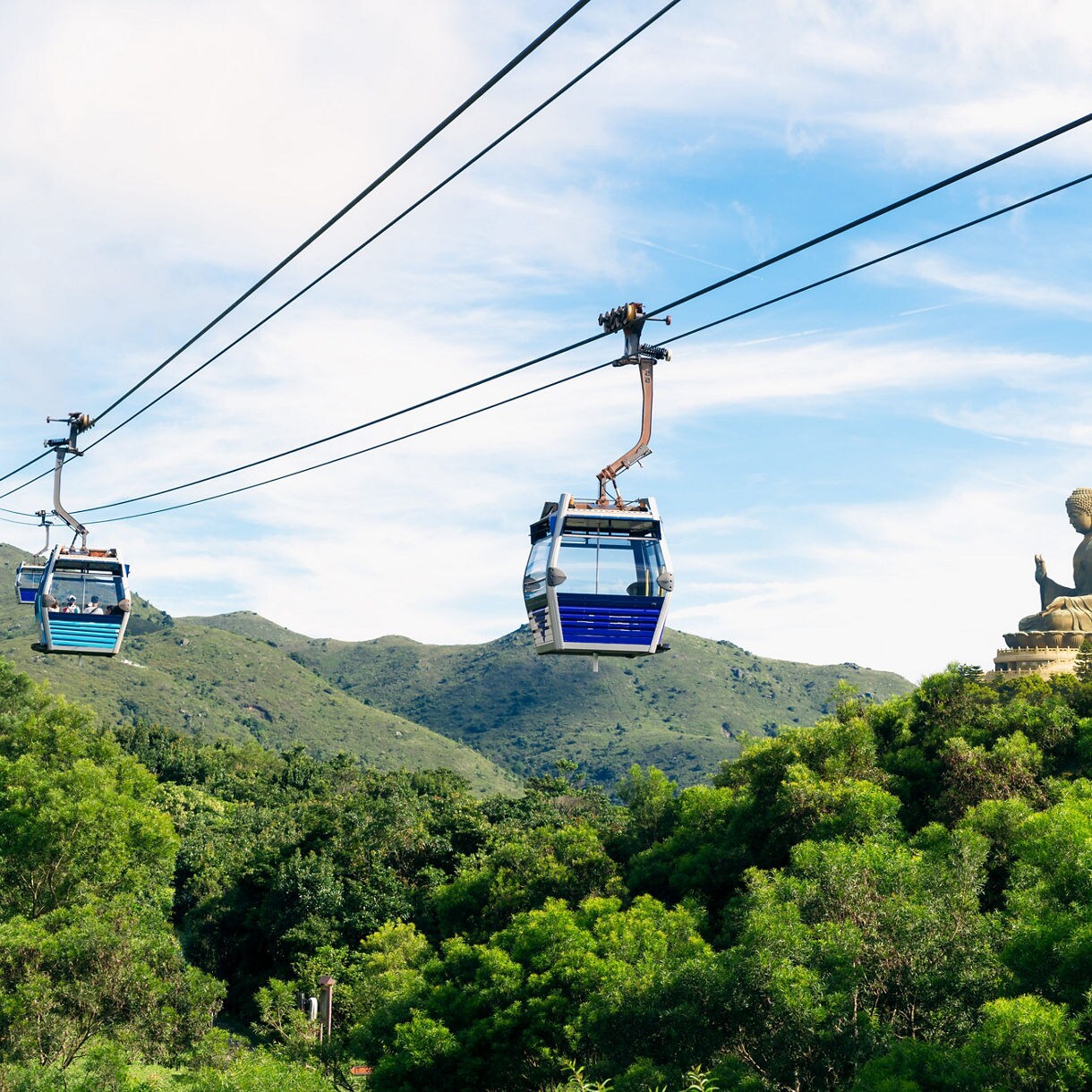Cable cars traveling over green hills toward the Tian Tan Buddha statue on Lantau Island under a bright blue sky.