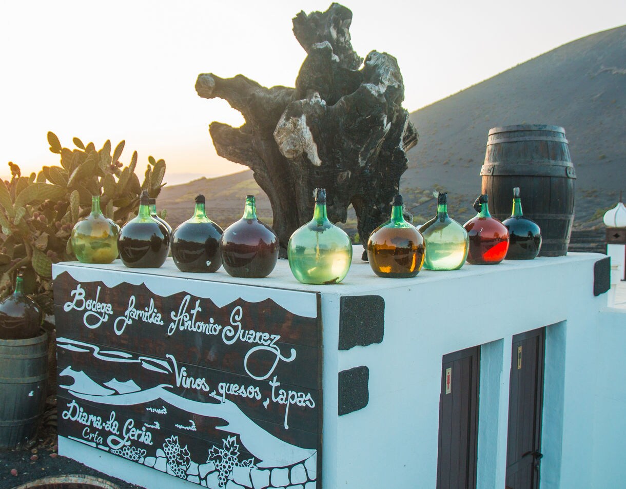 Row of colorful glass wine jugs atop a bodega wall in Lanzarote with cacti, a barrel, and volcanic hills in the background.