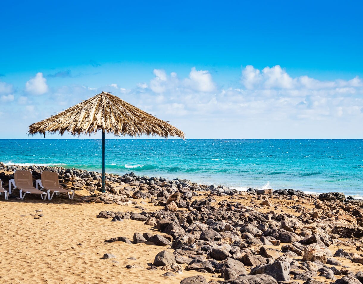 Sandy beach with volcanic rocks, a thatched umbrella, and lounge chairs overlooking the bright blue Atlantic Ocean in Lanzarote.