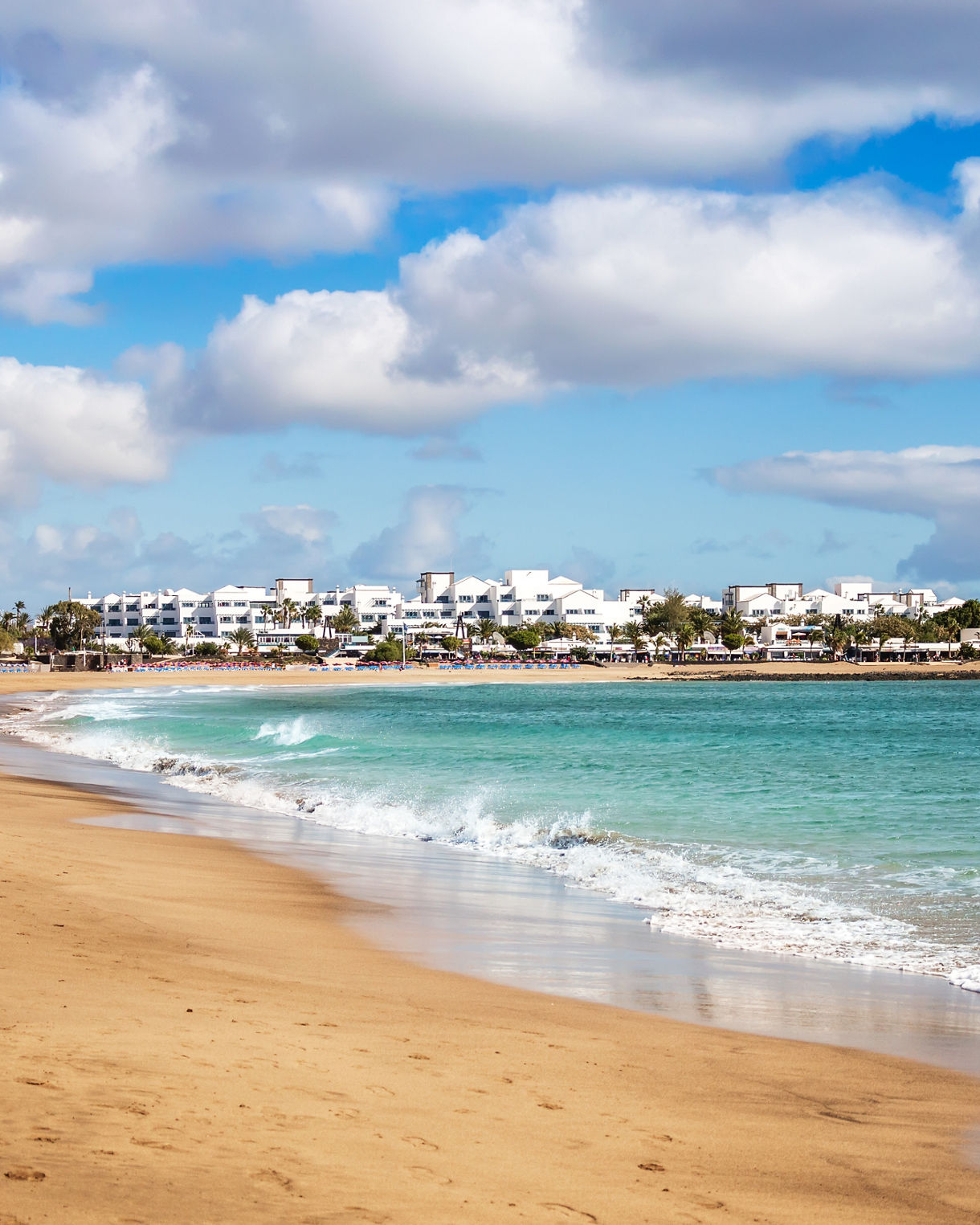 Wide sandy beach with gentle turquoise waves and whitewashed buildings of Puerto del Carmen under a bright blue sky with scattered clouds.
