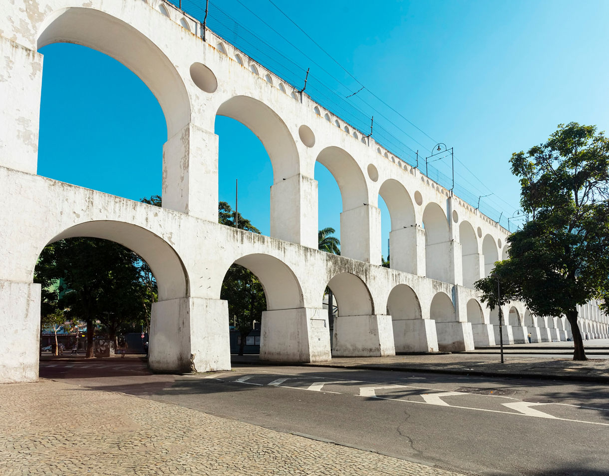 View of the white Lapa Arches in Rio de Janeiro, showing tall repeating stone arches, a clear blue sky and a few people walking through the open plaza nearby.