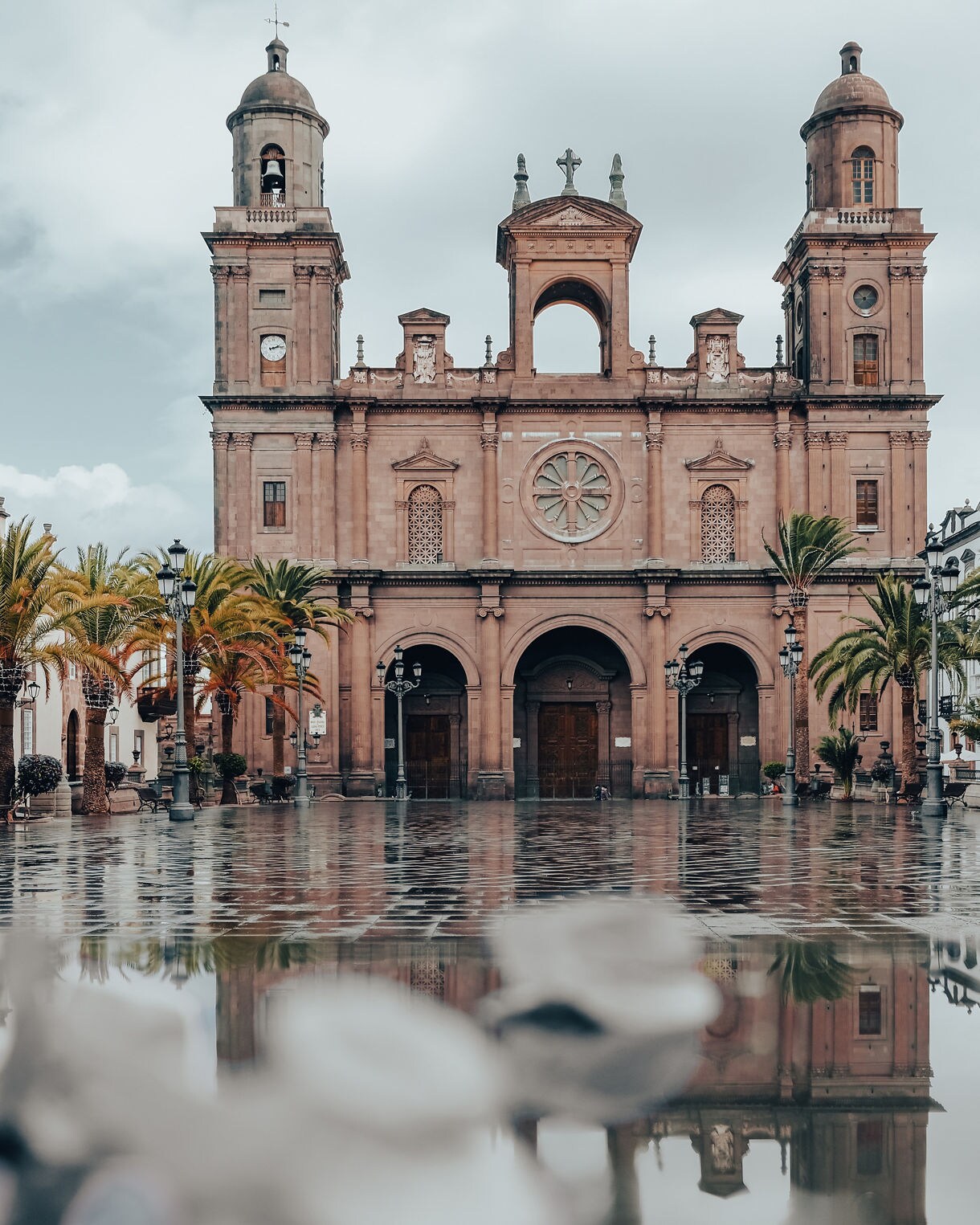 Front view of Las Palmas Cathedral in Gran Canaria, featuring twin bell towers, a rose window and arched entrances, reflected in puddles on the stone square.