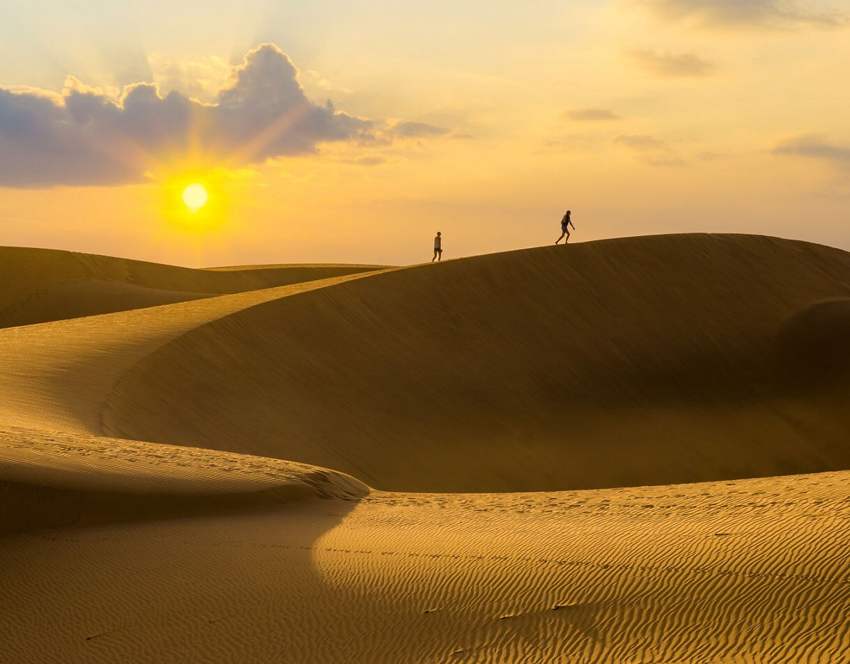 Silhouettes of two people walking along the crest of the Maspalomas Dunes in Gran Canaria at sunset, with rippled sand glowing under the warm light.