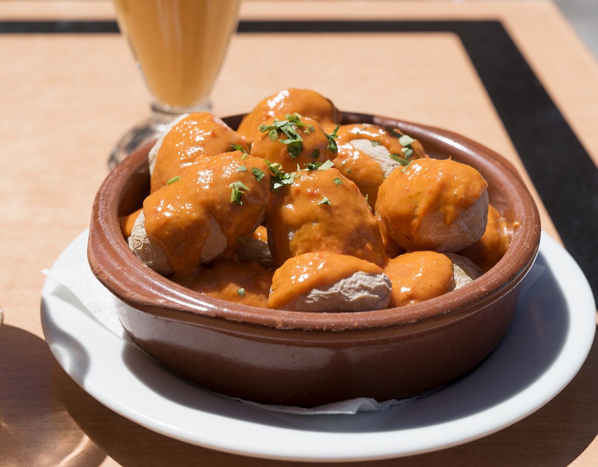 Clay dish filled with papas arrugadas, small wrinkled potatoes covered in red mojo sauce and garnished with chopped herbs, served on a white plate.