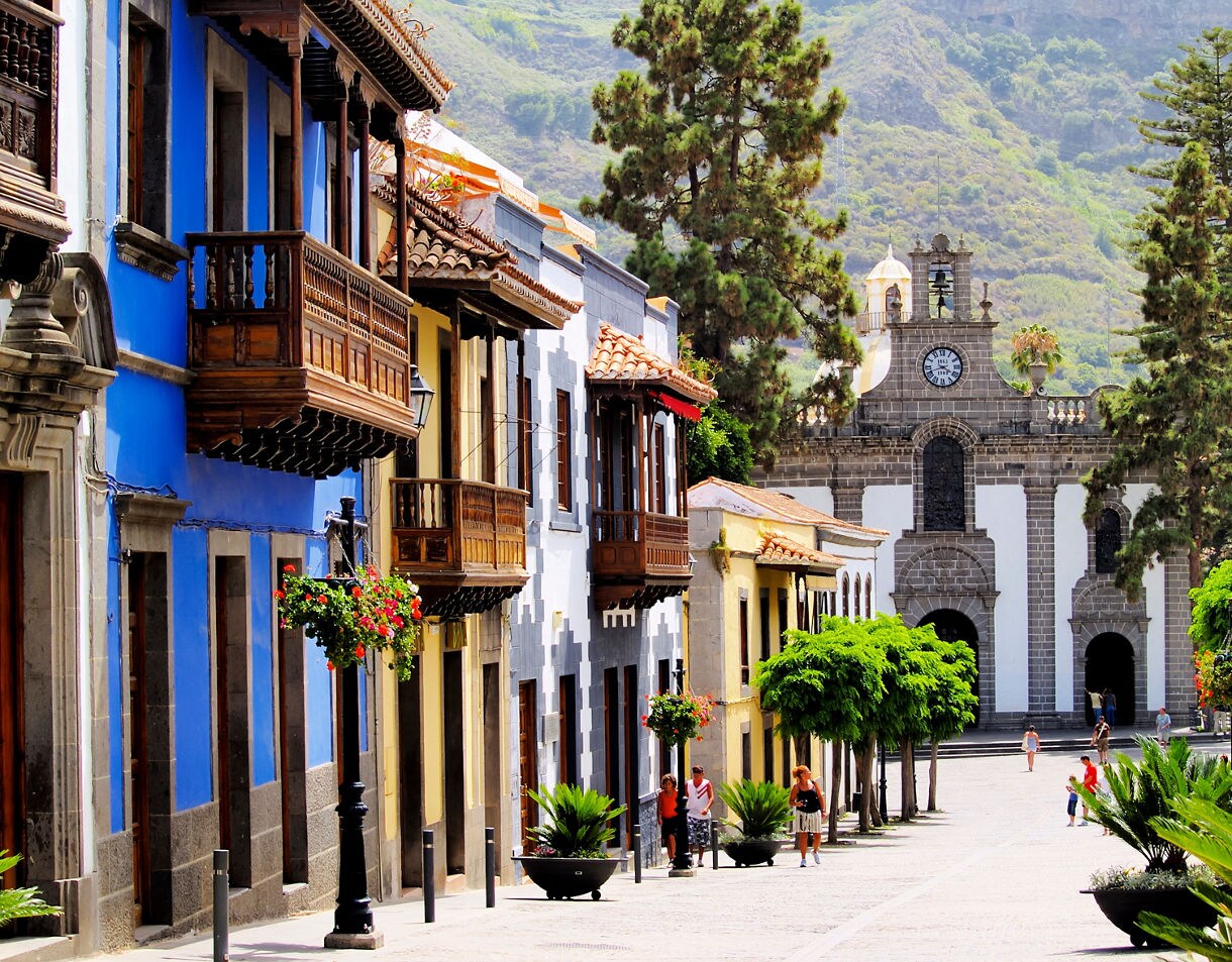 Brightly painted houses with carved wooden balconies line the streets of Teror, Gran Canaria, with the Basilica of Our Lady of the Pine framed by trees in the background.