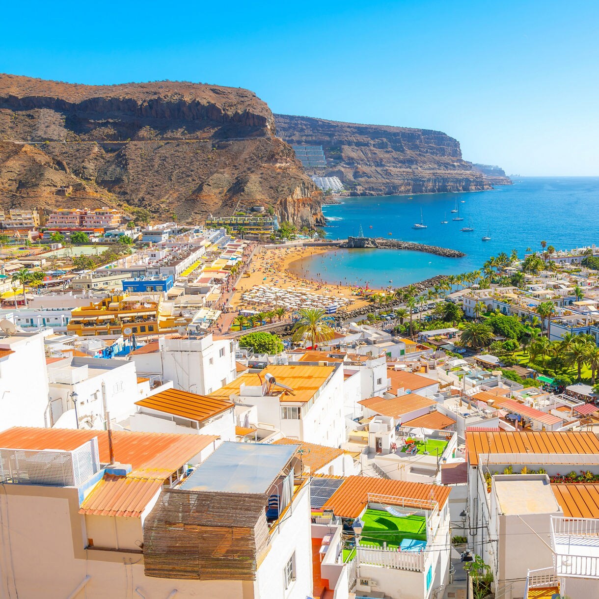 Aerial view of Puerto de Mogán in Gran Canaria with whitewashed houses, terracotta rooftops, a sandy beach filled with umbrellas and the marina lined with boats beside rugged coastal cliffs.