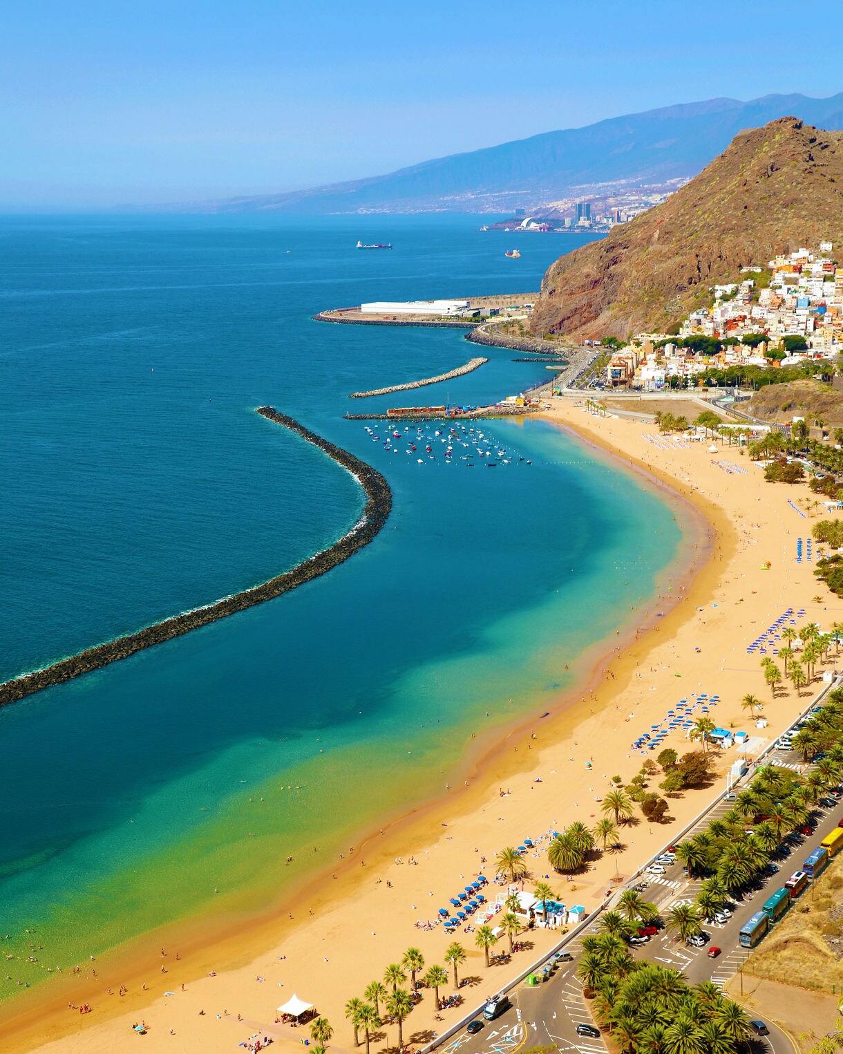 Aerial view of Las Teresitas Beach in Tenerife, showing golden sand, clear blue water, palm trees and colorful umbrellas along the shoreline.