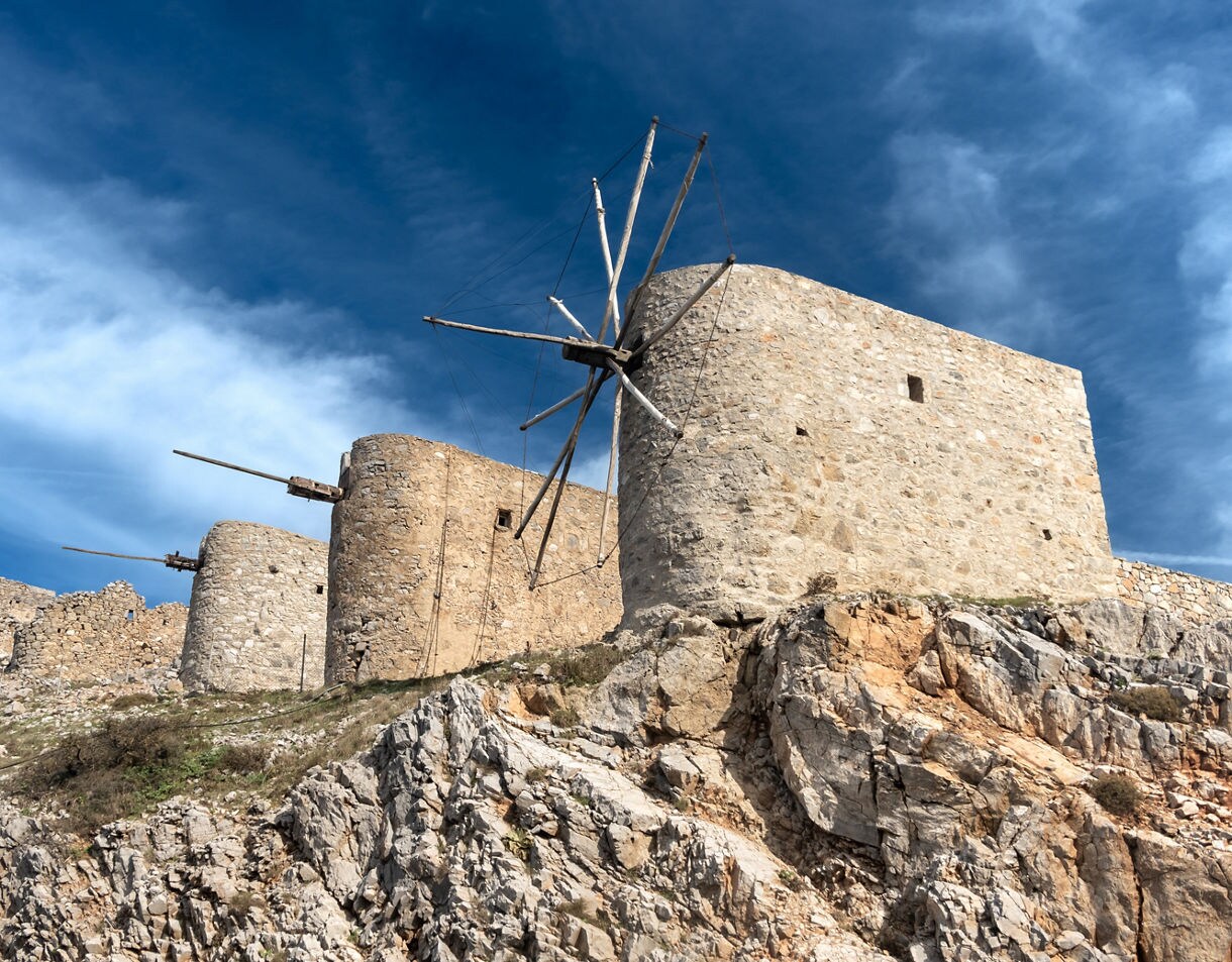 Row of old circular stone windmills perched on a rocky hillside in the Lassithi Plateau of Crete, featuring weathered masonry, wooden sail frames and a dramatic blue sky overhead.