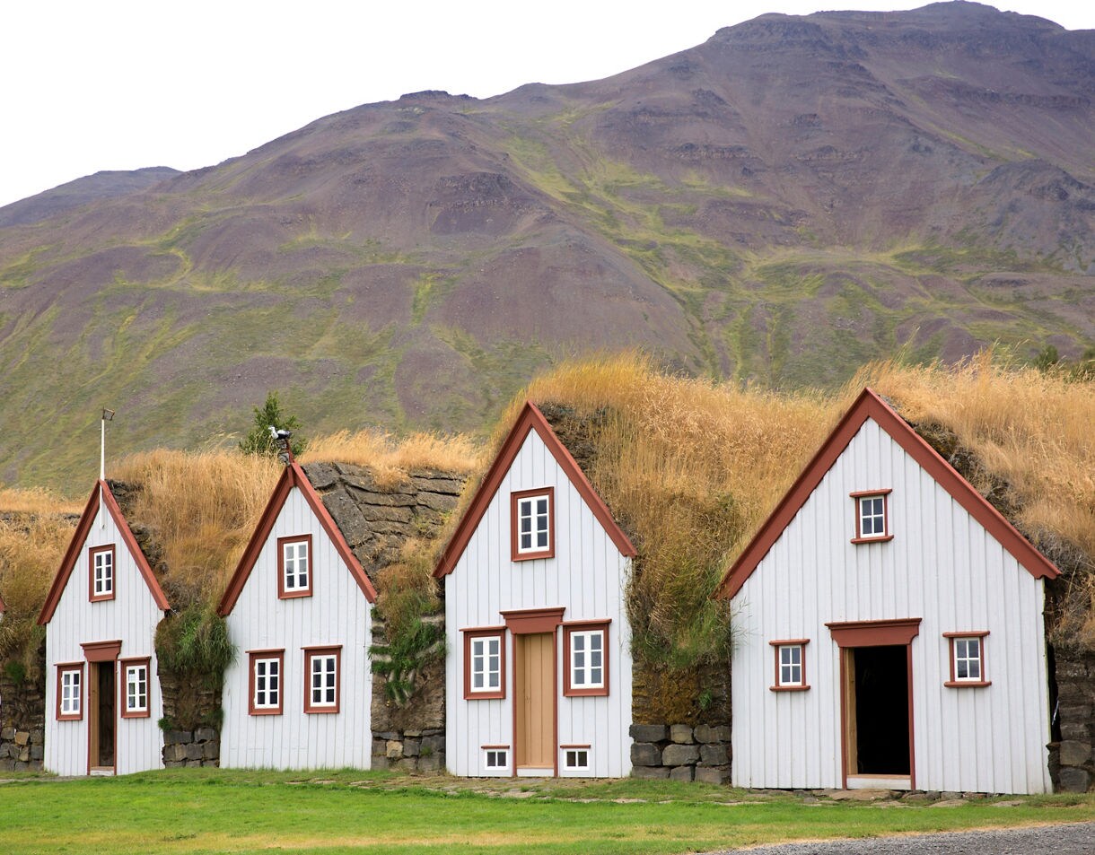 Row of white wooden turf houses with red trim at Laufás Folk Museum in Iceland, built into grassy mounds at the base of a rocky hillside.