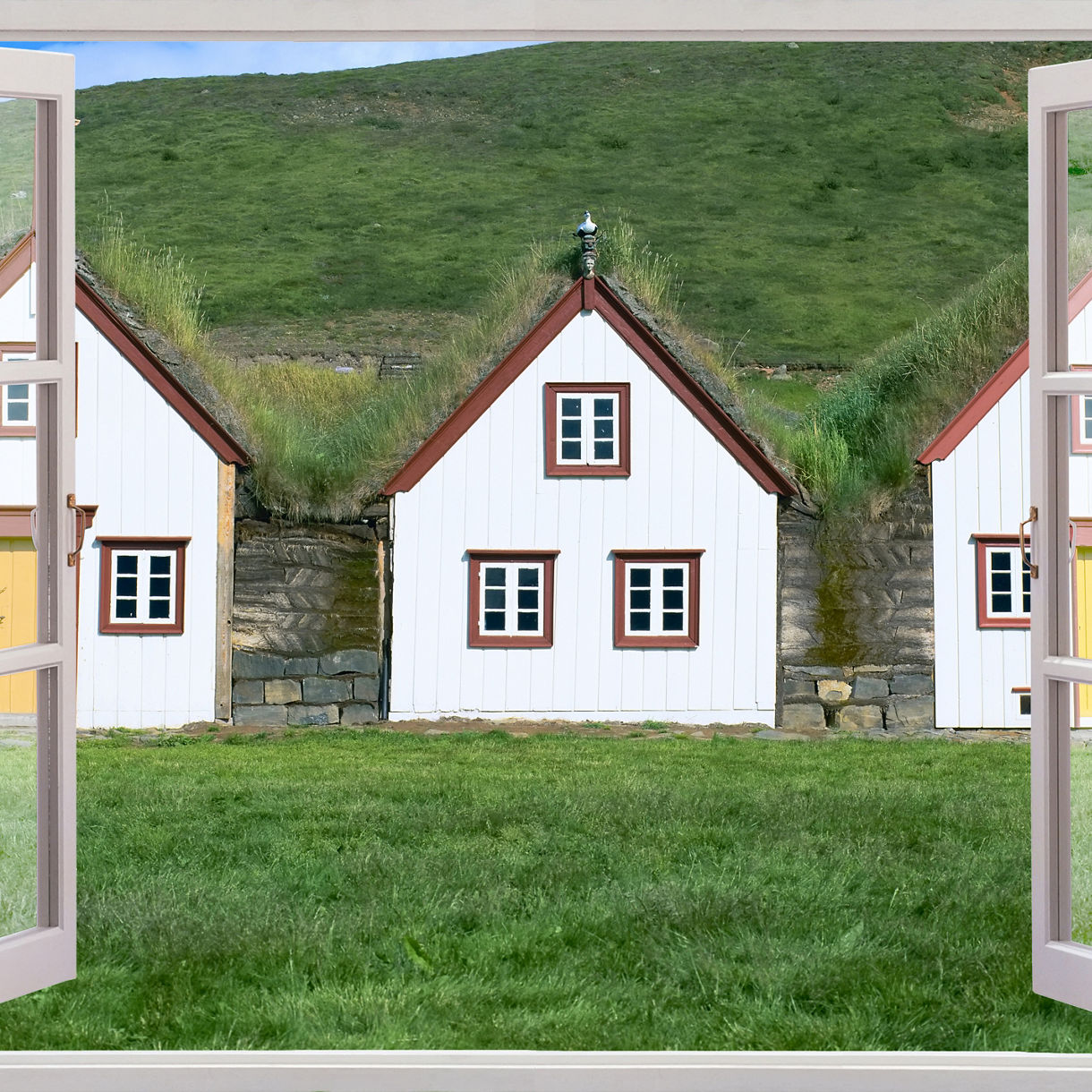 View through an open window of traditional white turf houses with grass-covered roofs at Laufás Folk Museum in Iceland, set against a green hillside.