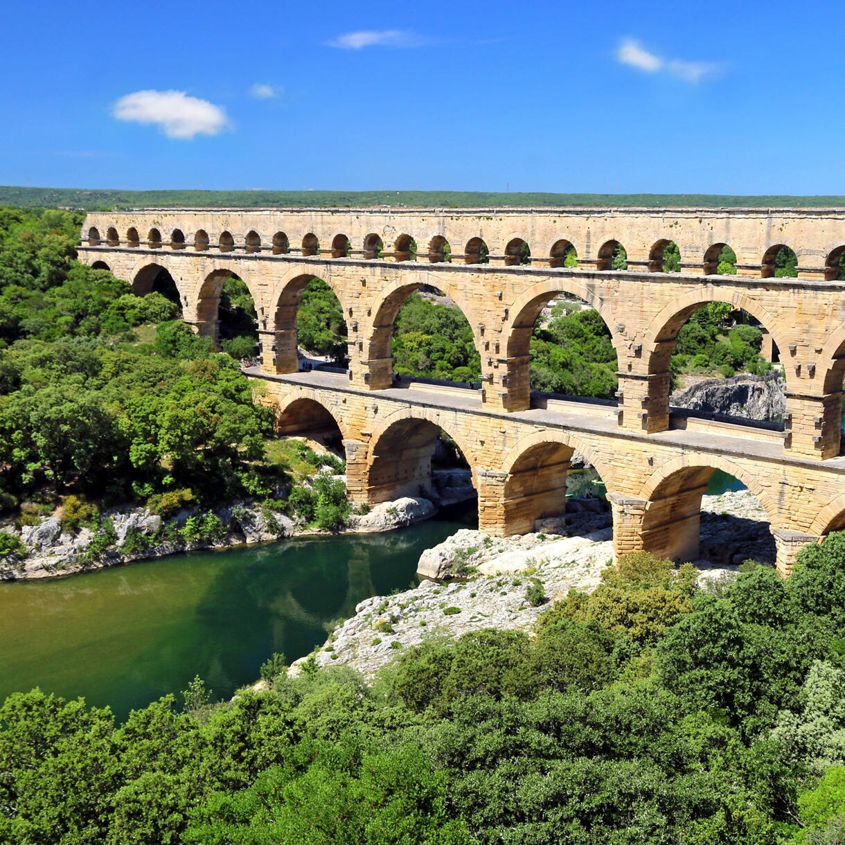 Ancient three-tiered stone aqueduct, the Pont du Gard, spanning the Gardon River with lush green hills surrounding it.