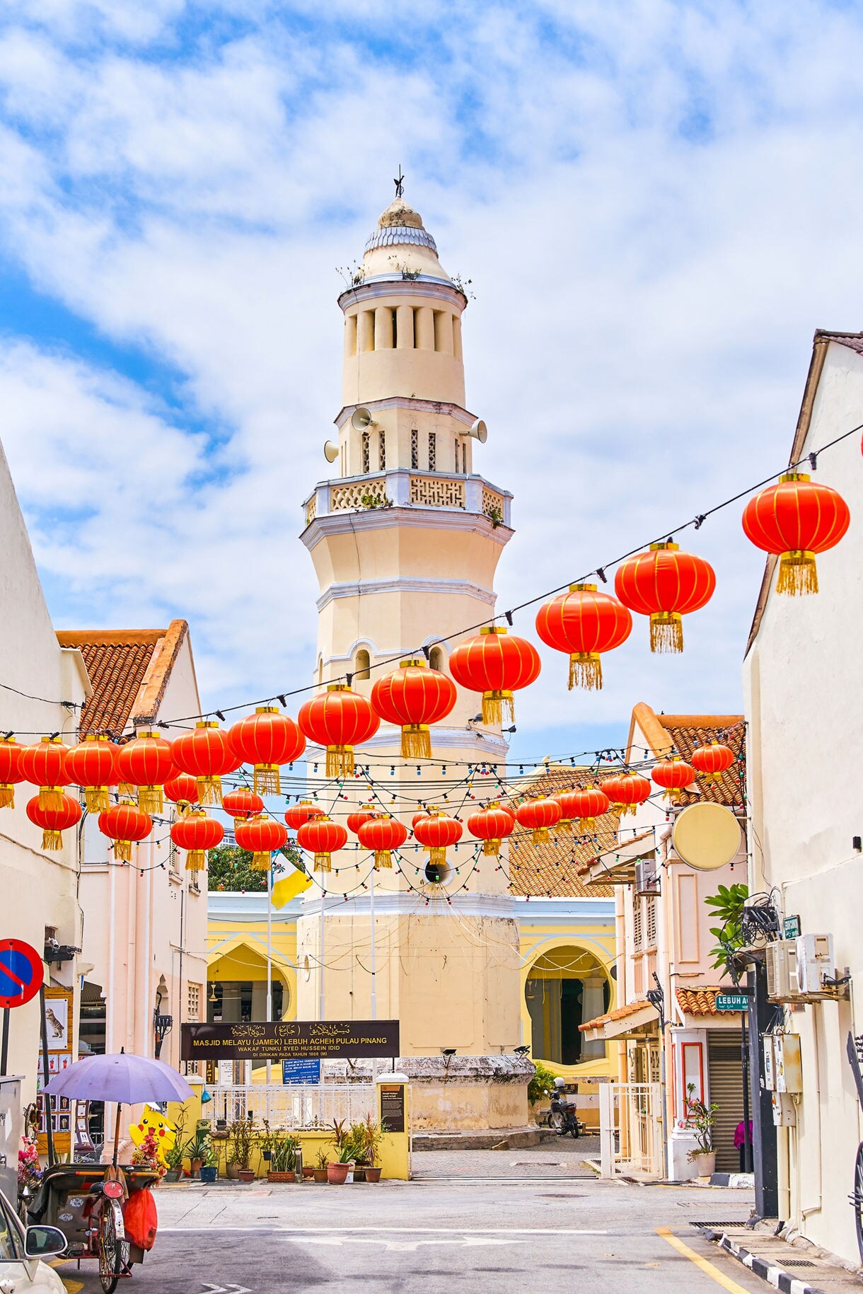 Tall cream-colored minaret of Acheen Street Mosque in George Town, Penang, with rows of red Chinese lanterns hanging across the street.