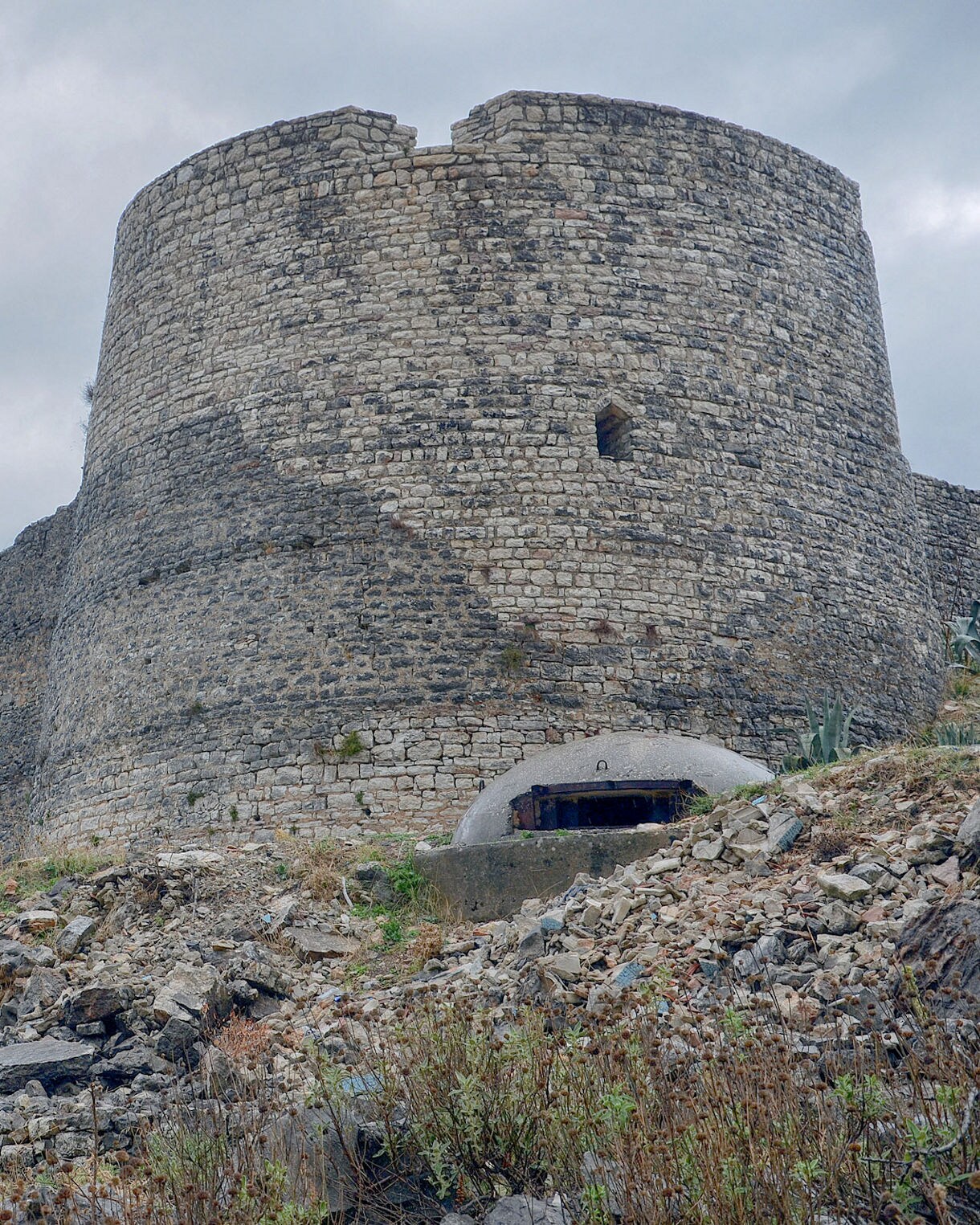 Stone walls and a large round tower of Lëkurësi Castle rise above rocky ground and dry vegetation under an overcast sky in southern Albania.